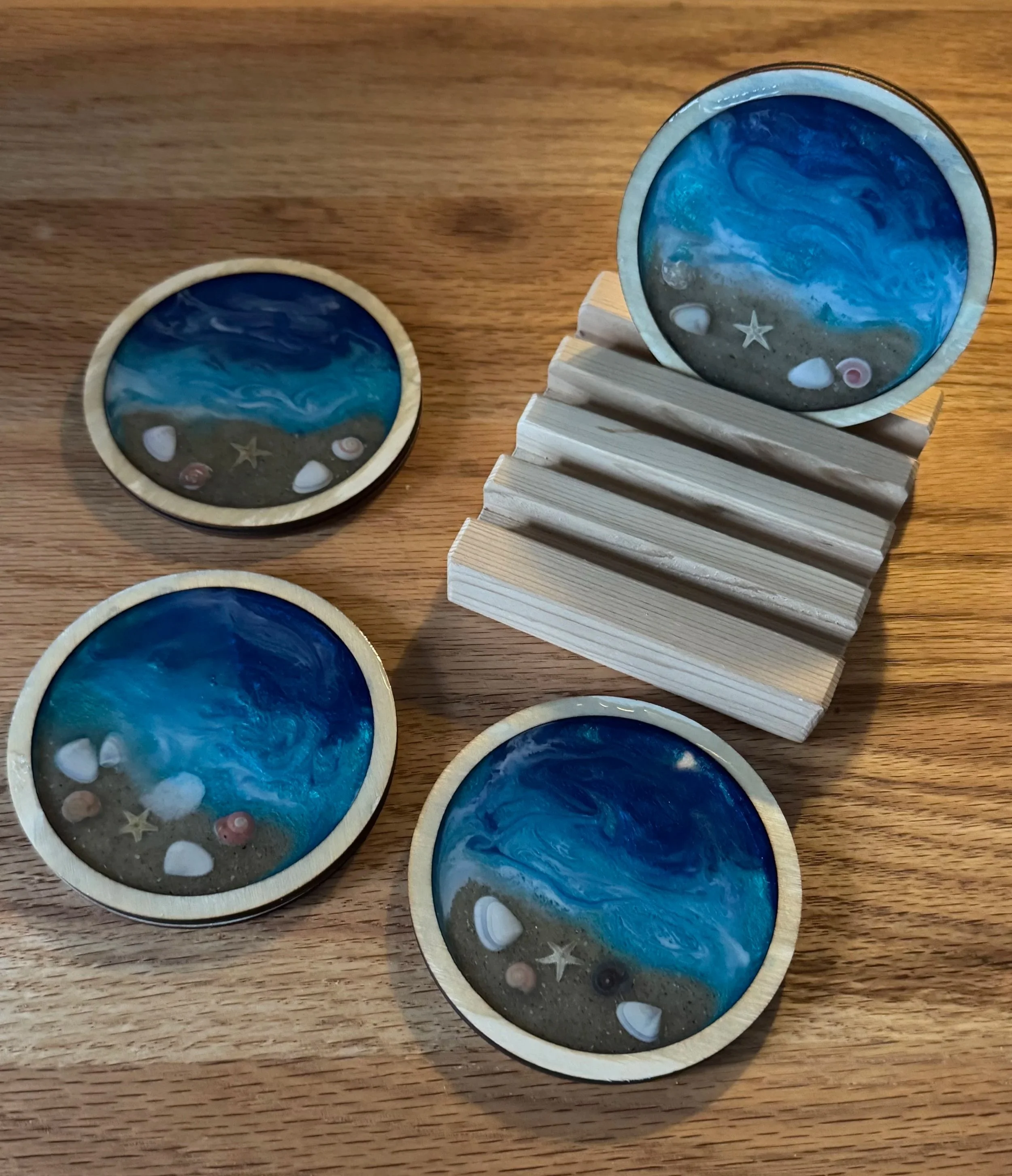 Multiple round coasters with ocean and beach designs, featuring seashells, starfish, and sand, displayed on a wooden surface with a wooden slatted coaster holder.