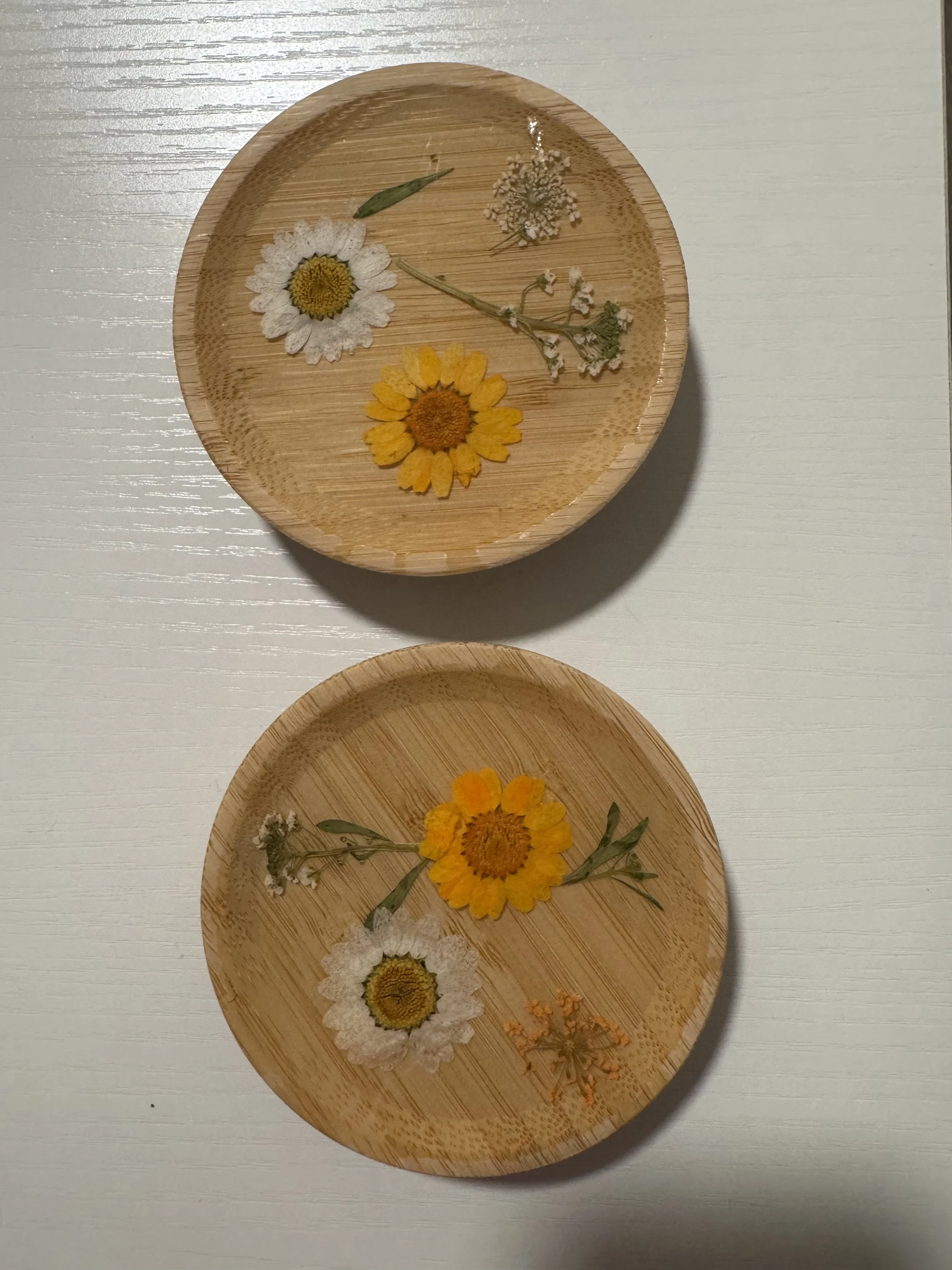 Two round wooden dishes on a white surface, each containing dried flowers including daisies, small white flowers, and greenery.