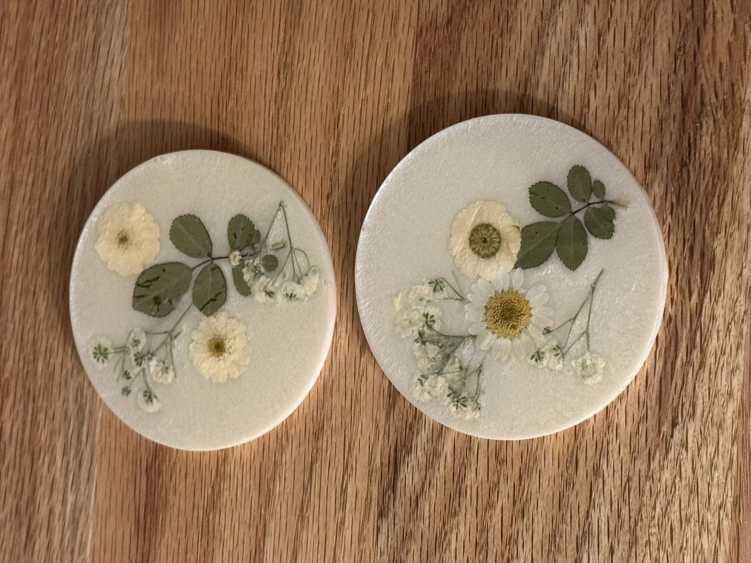 Two white ceramic plates with pressed flowers and green leaves, placed on a wooden surface.
