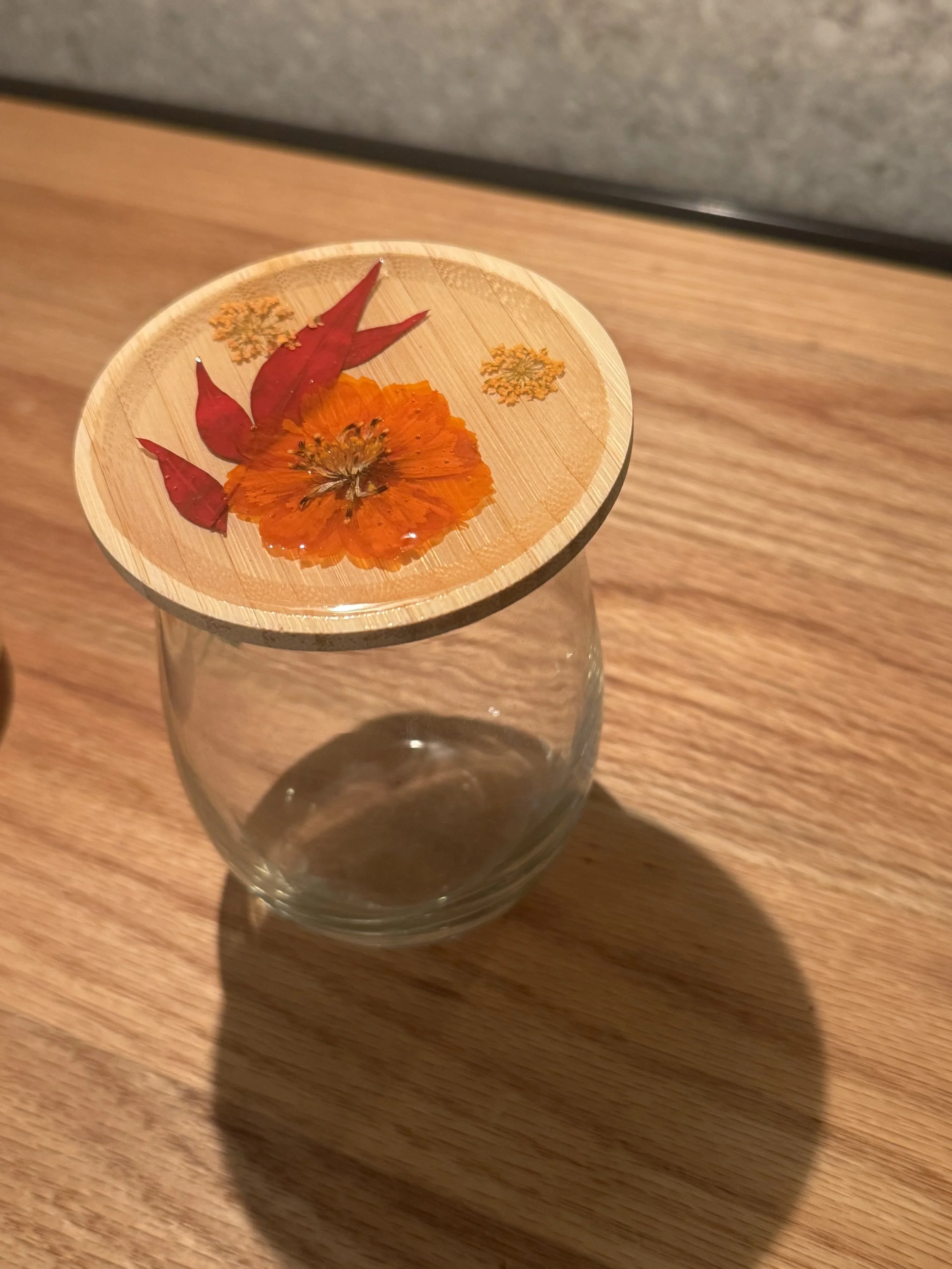 A glass with a wooden lid decorated with dried flowers and red leaves, sitting on a wooden surface.