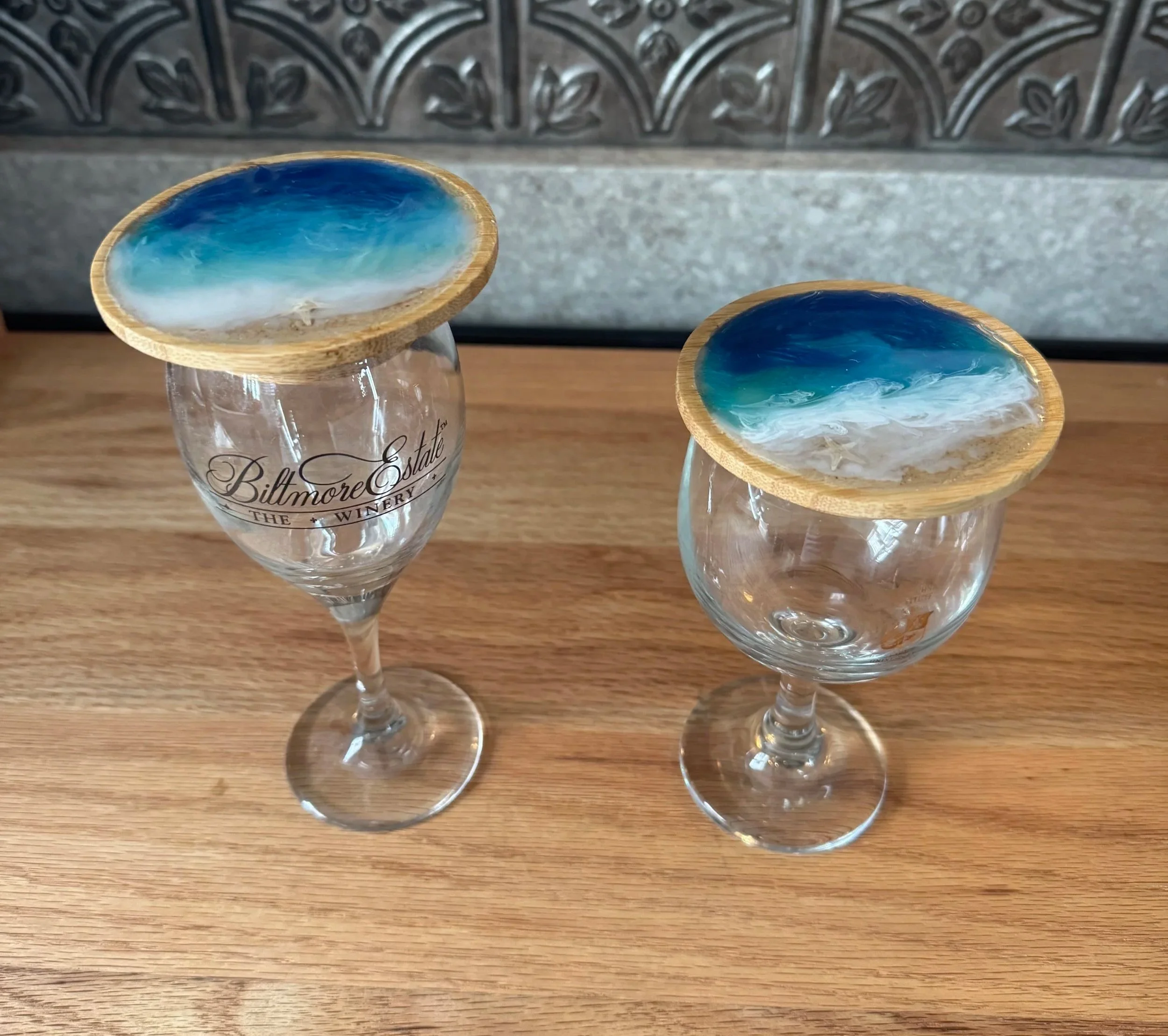 Two glasses with seashell-shaped resin tops featuring blue and white ocean wave designs, placed on a wooden surface with a decorative tile background.
