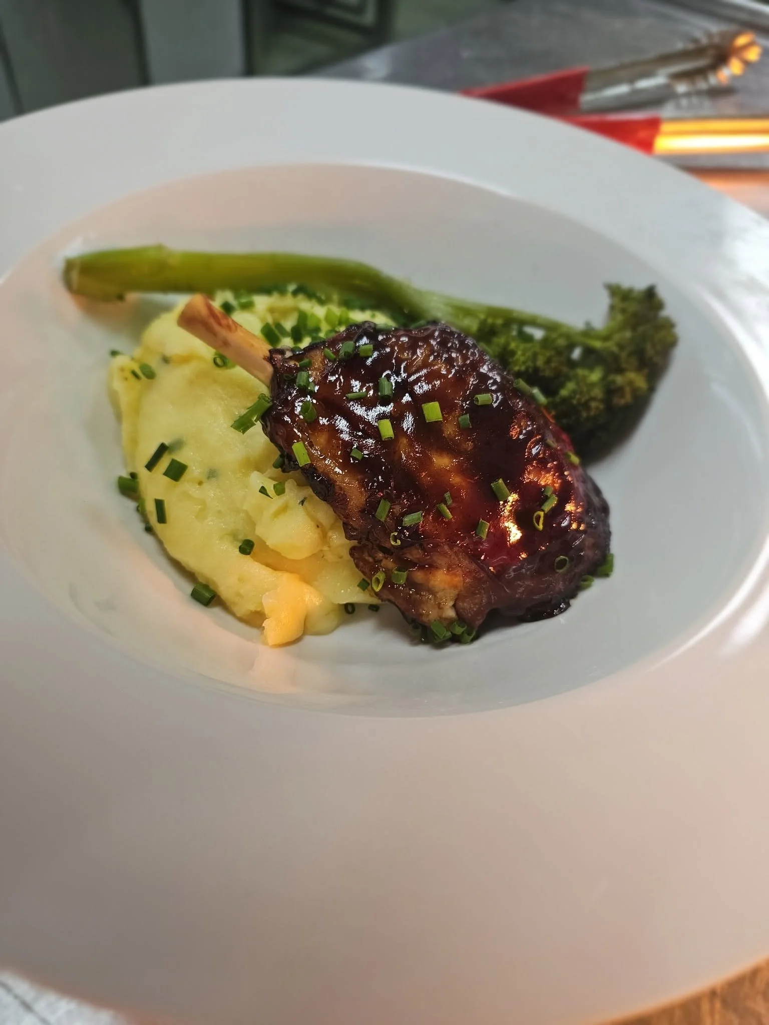 A white plate contains mashed potatoes topped with chopped chives, a green grilled asparagus spear, and a glazed meat or protein rib with a dark sauce, garnished with chopped chives.