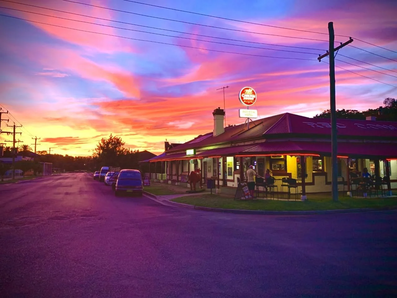 A restaurant/bar with a red neon roof and a lit sign, situated on a street during sunset. The sky is vibrant with shades of pink, orange, and purple. There are cars parked along the street and a few people sitting outside at tables.