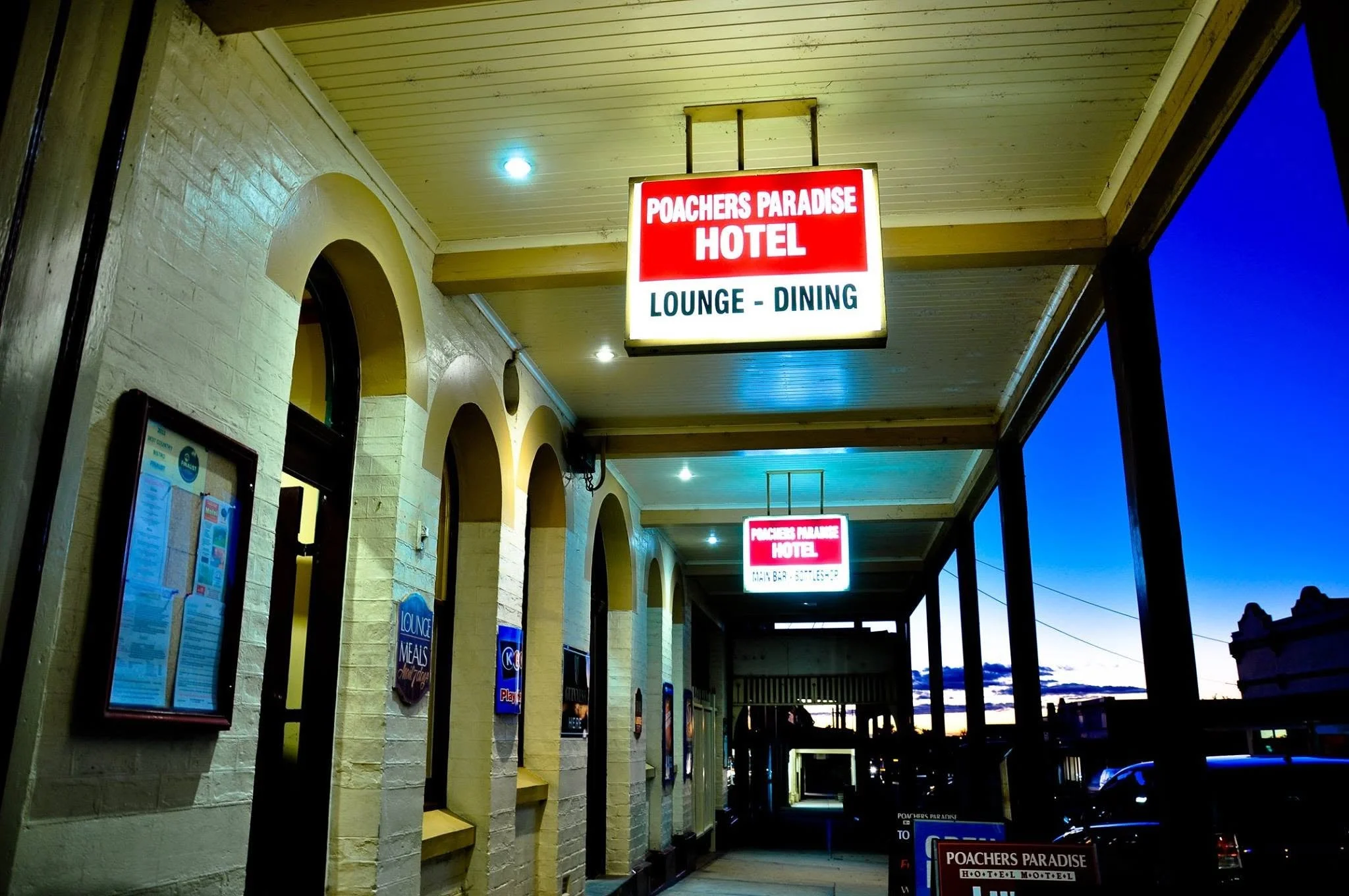 Signboard for Poachers Paradise Hotel with lounge and dining options at night, part of an outside corridor of a restaurant or hotel with illuminated signs on a building with arched windows.