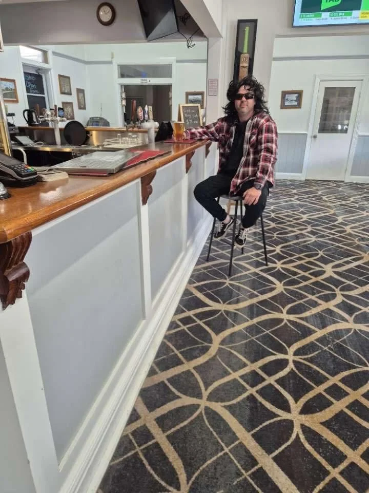 A man with curly hair wearing sunglasses, a red plaid shirt, and black pants is sitting on a barstool at a bar area in a restaurant or pub. He is holding a glass of beer.