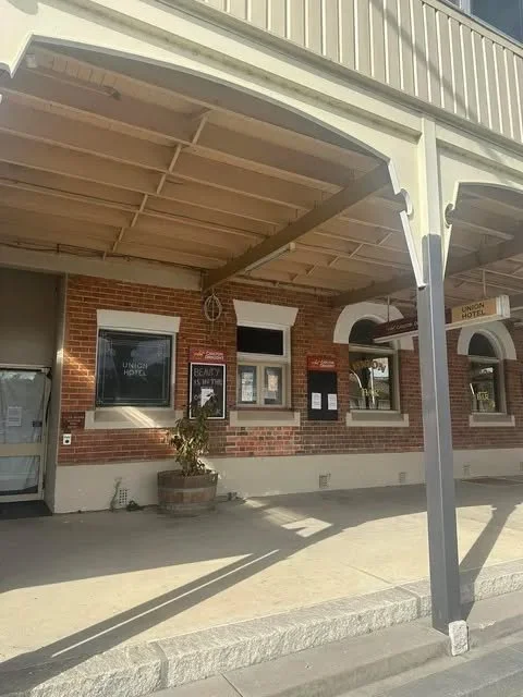 Side view of a brick building with a covered sidewalk, windows displaying posters, and a potted plant.