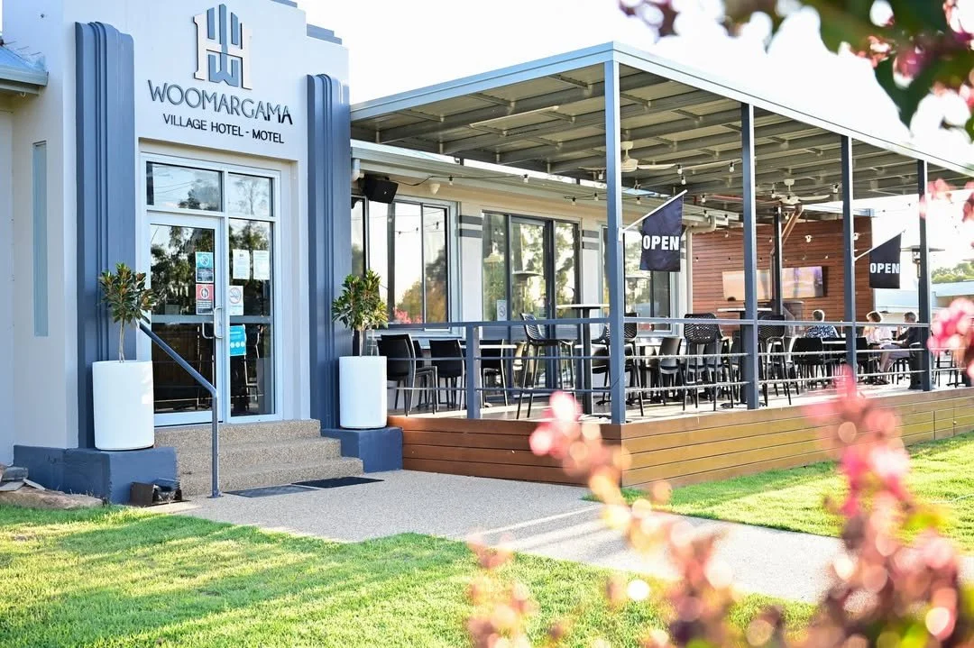 Exterior view of Woomarga Village Hotel-Motel with a porch area, outdoor seating, and open signs, surrounded by grass and pink flowering plants.