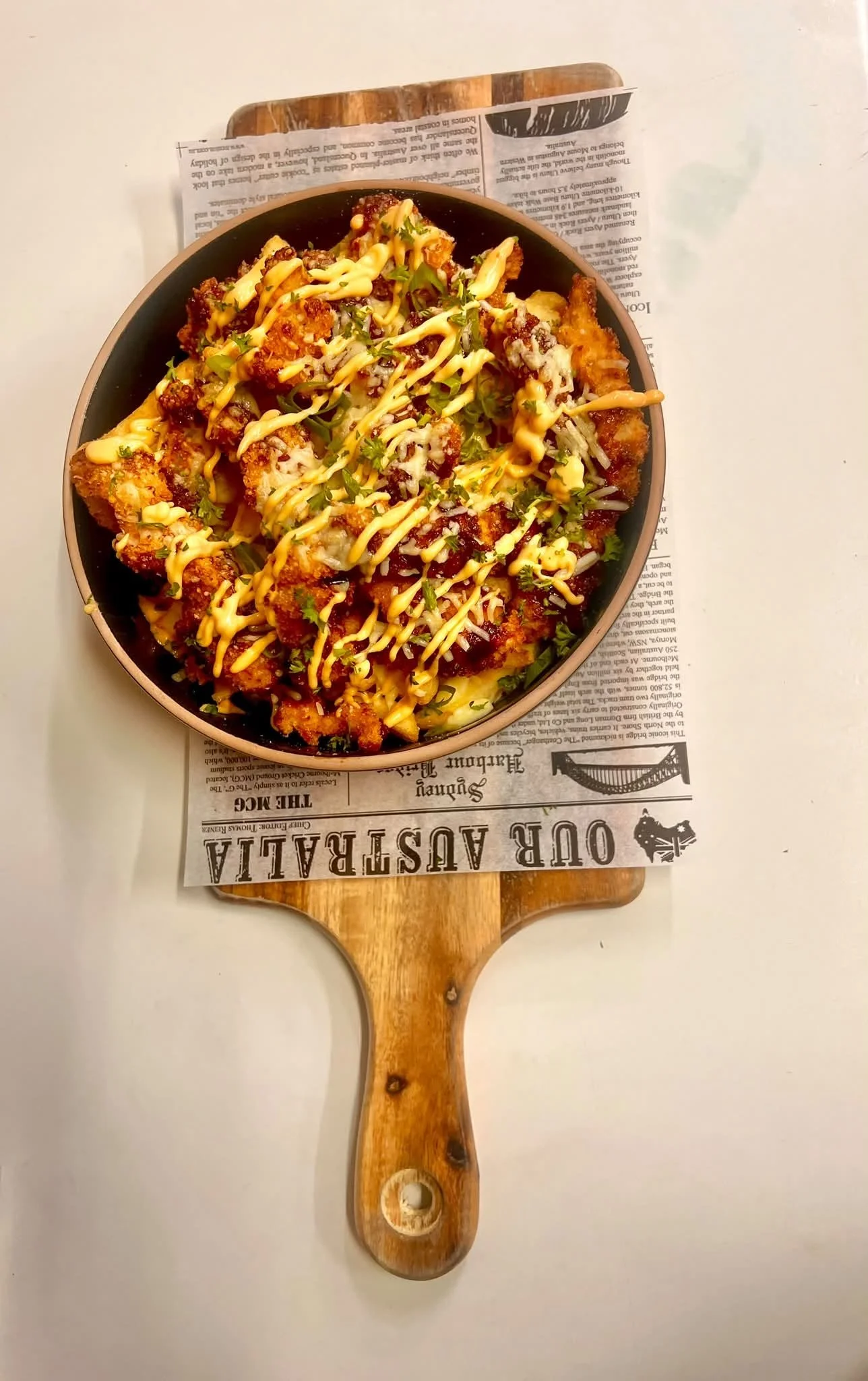 A bowl of loaded nachos topped with cheese, sauce, and herbs, placed on a wooden serving board with a paper underneath that has vintage-style print.