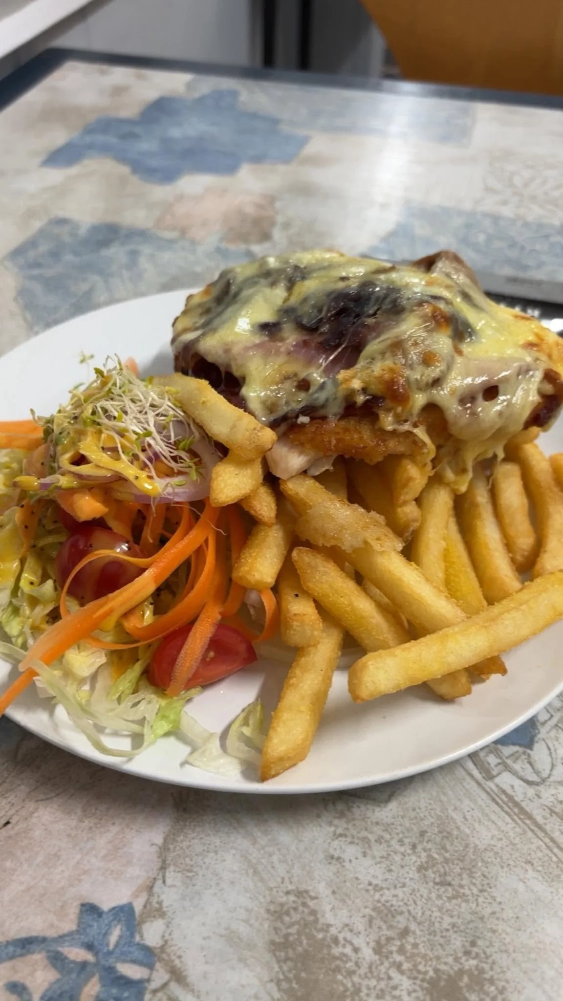 Plate of French fries, salad with cherry tomatoes, shredded carrots, and sprouts, topped with cheese-covered breaded chicken.