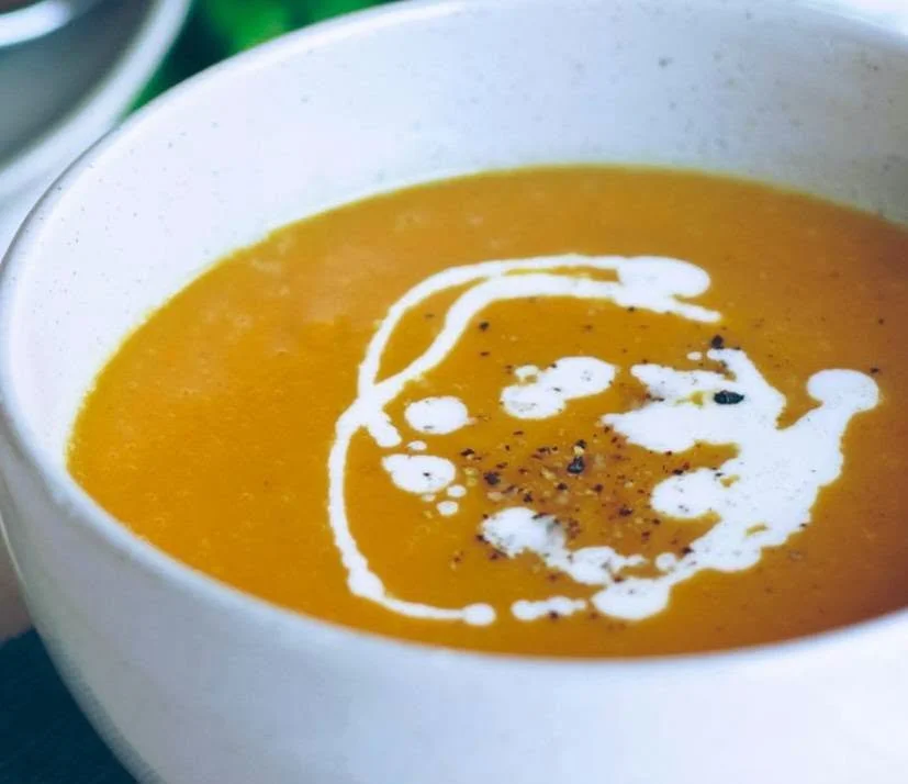 A white bowl filled with orange soup, topped with a swirl of white cream and sprinkled with black pepper.