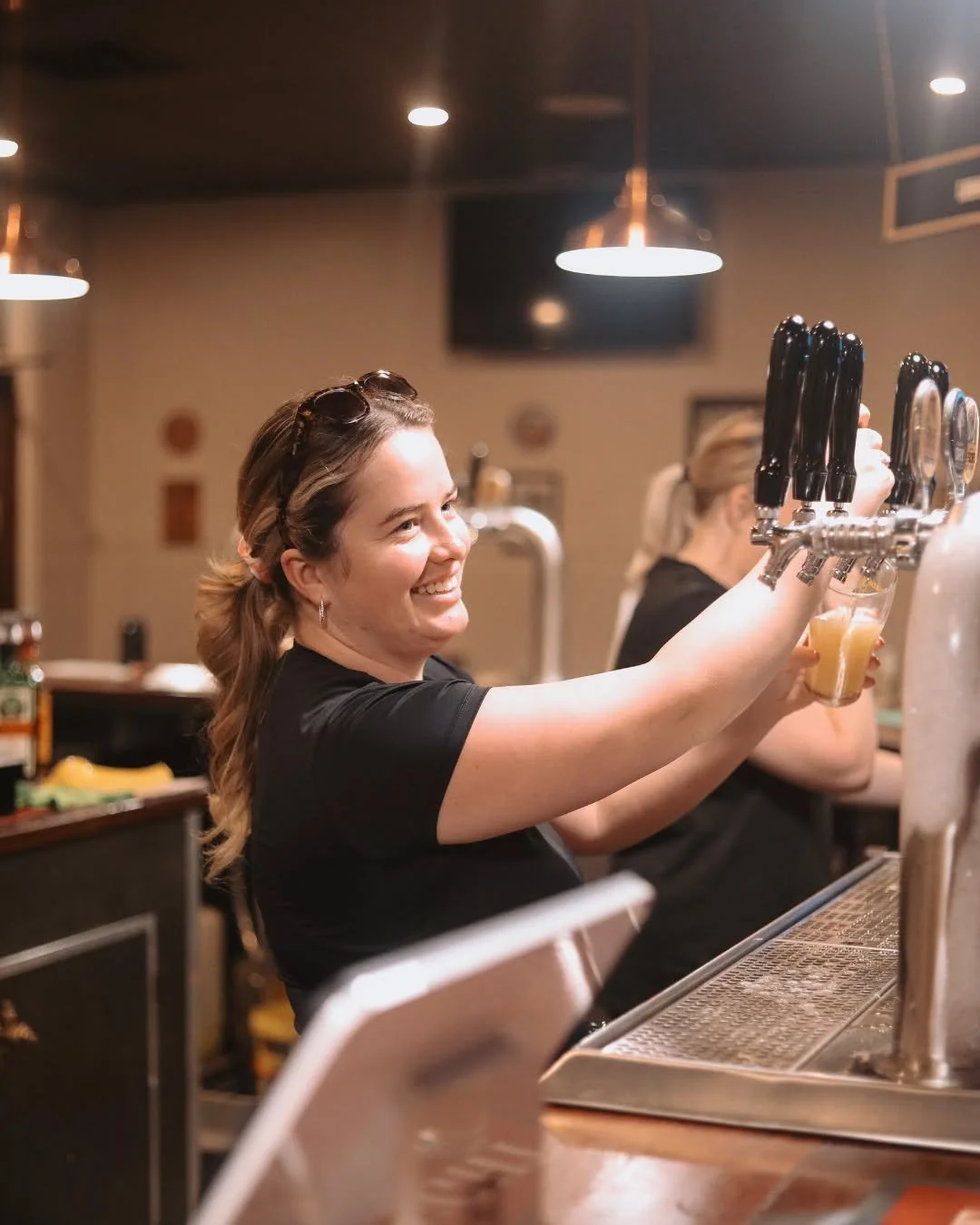 A woman smiling while pouring beer from tap handles at a bar.