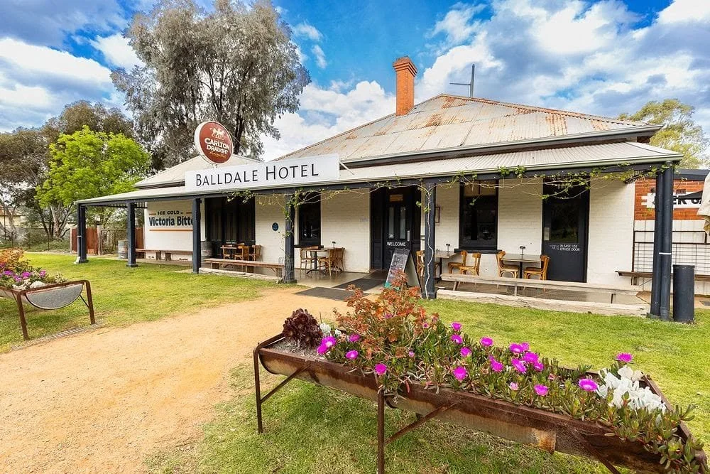 Front view of the Balldale Hotel, a historical building with a white brick exterior, black window frames, and a metal roof, surrounded by greenery and flower beds, under a partly cloudy sky.