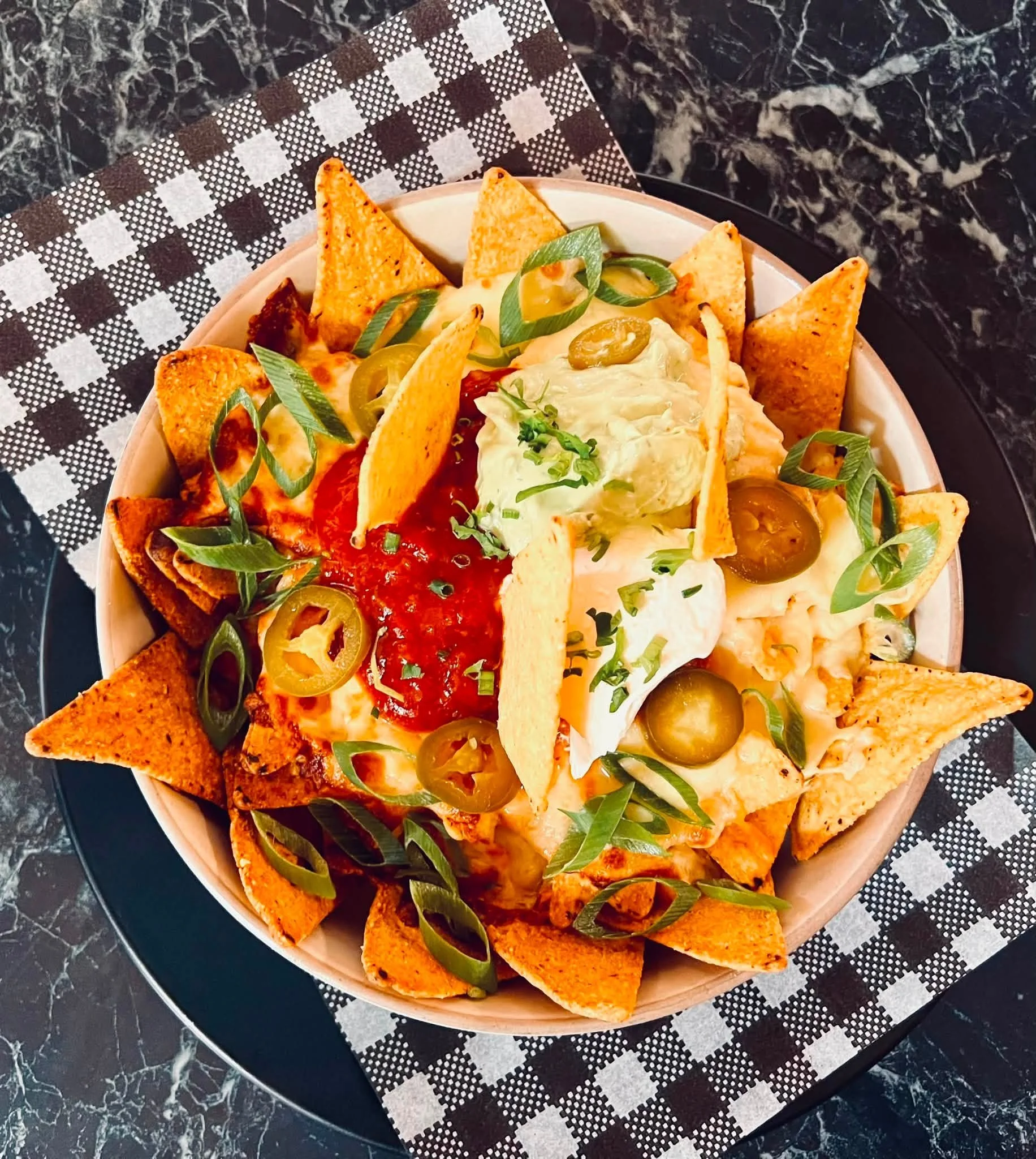 A bowl of nachos topped with cheese, jalapenos, diced tomatoes, shredded lettuce, and melted cheese, served on a black marble table with a black and white checkered napkin underneath.