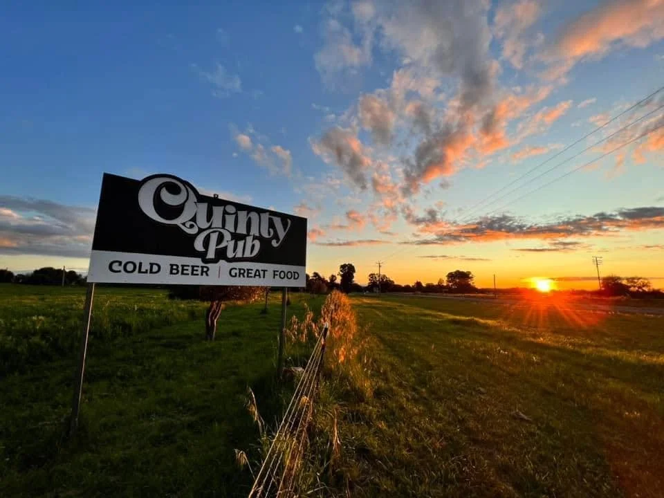 Sunset over a grassy field with a sign for Quiny Pub advertising cold beer and great food.