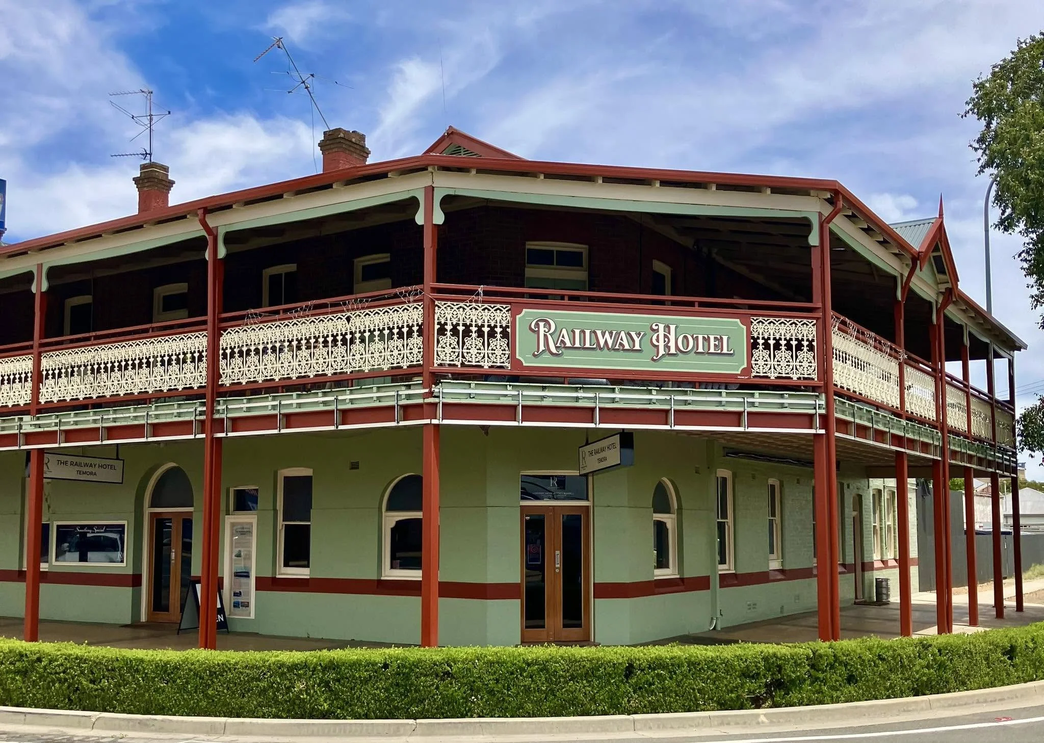 Two-story historic building with a sign that reads 'Railway Hotel'. The building has a green lower level, decorative white railing, and a red roof with multiple chimneys. The sky is partly cloudy with blue patches, and there is a tree on the right si