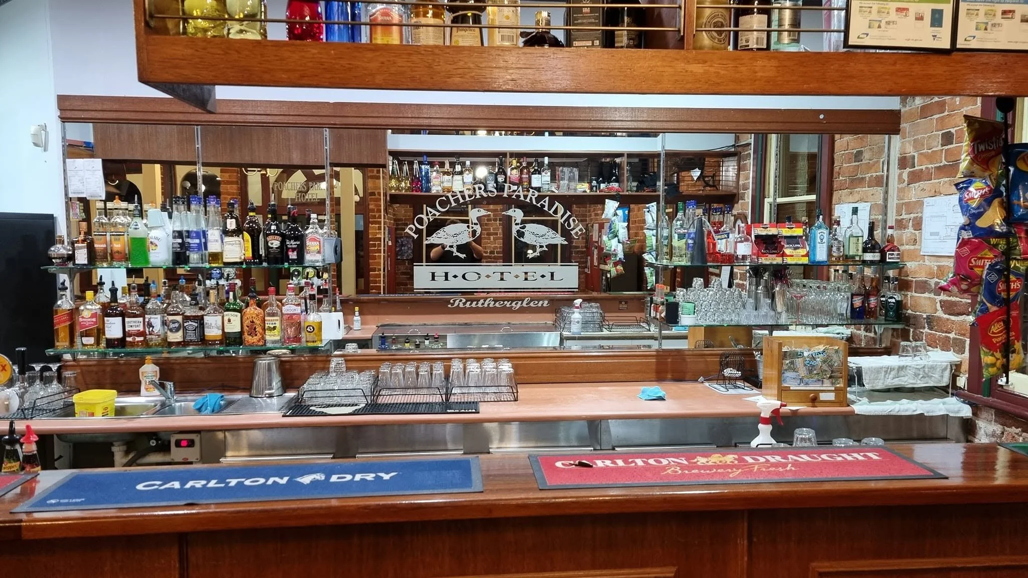 Bar area at Poacher's Paradise Hotel in Rutherglen, featuring bottles of alcohol on glass shelves, glasses, and a mirror with the hotel's name and two geese logo.
