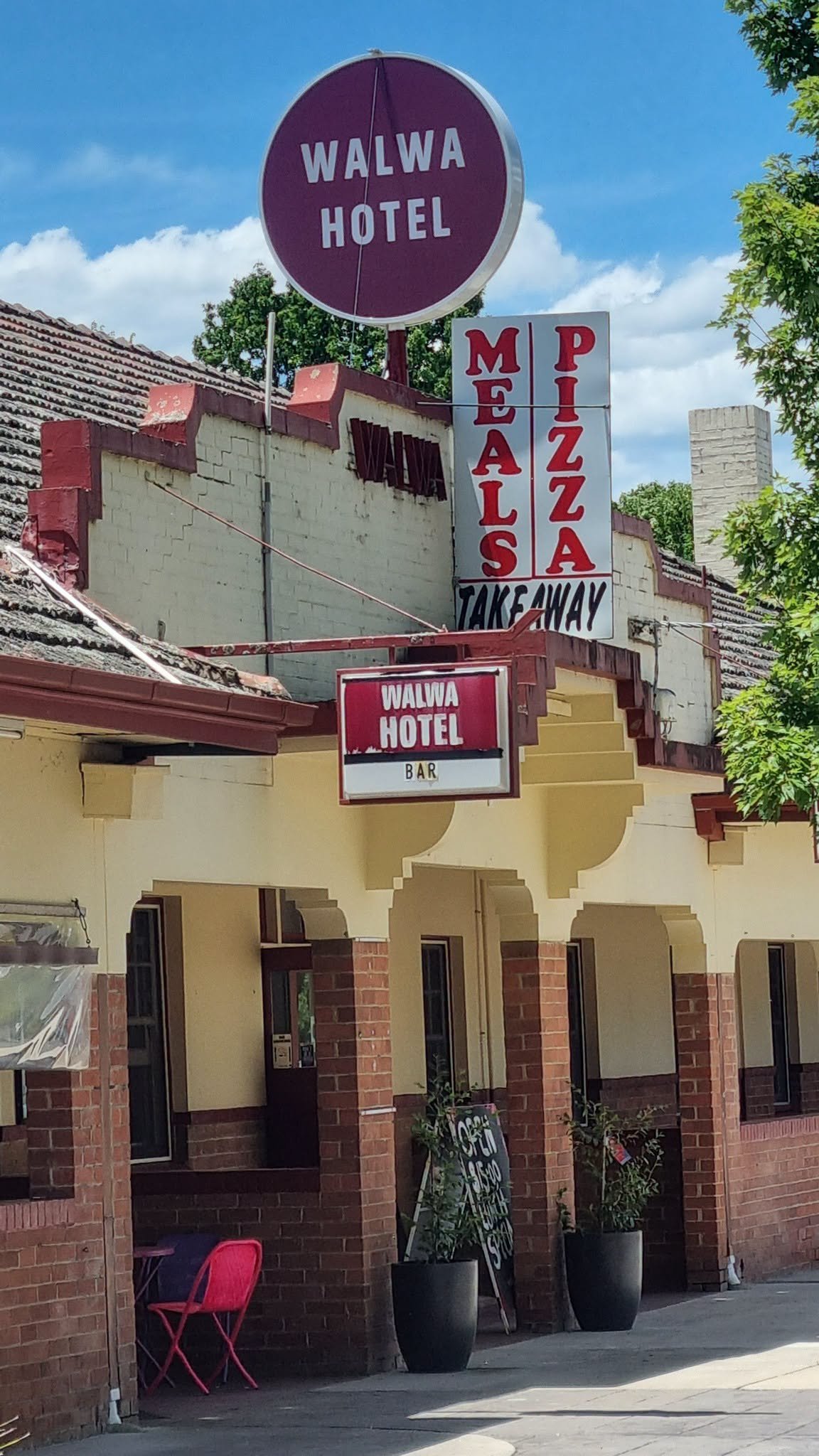 Exterior of Walwa Hotel, a building with a brick facade, multiple signs for the hotel, pizza, and takeaway, and potted plants outside on a sunny day.