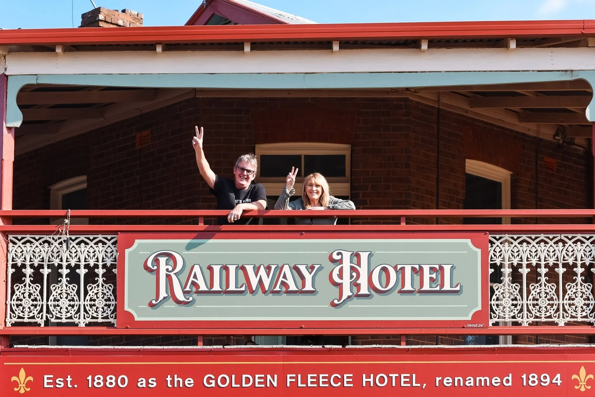 Two smiling people waving from a balcony of the Railway Hotel, a historic building with brick walls, decorative white ironwork, and a large sign that states it was established in 1880.