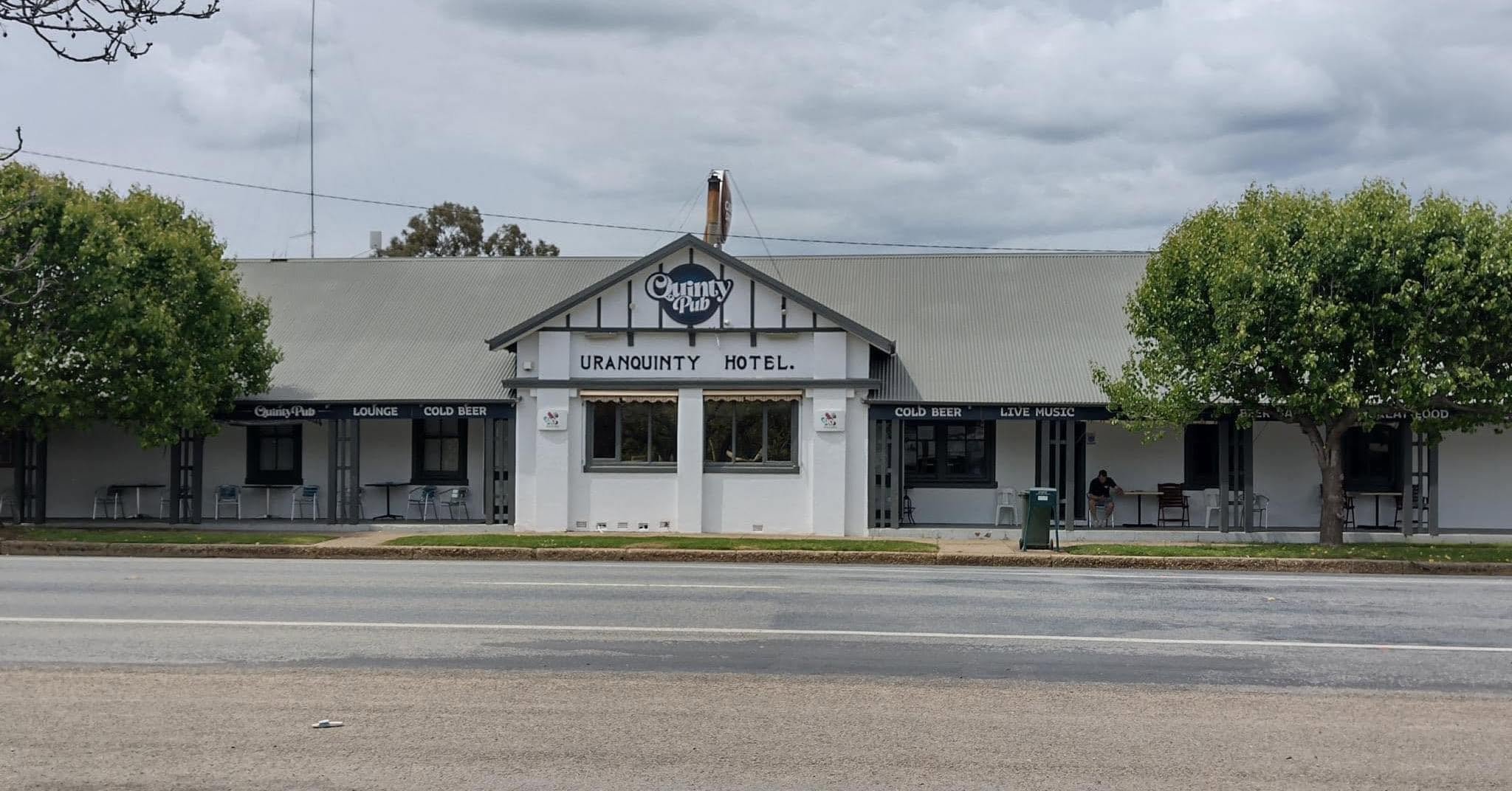 Front view of the Uranquinty Hotel, a single-story building painted white with a grey roof. A sign on the building reads 'Uranquinty Hotel' and 'Uranquinty Pub.' The building features an outdoor seating area with chairs and tables, and signage indica