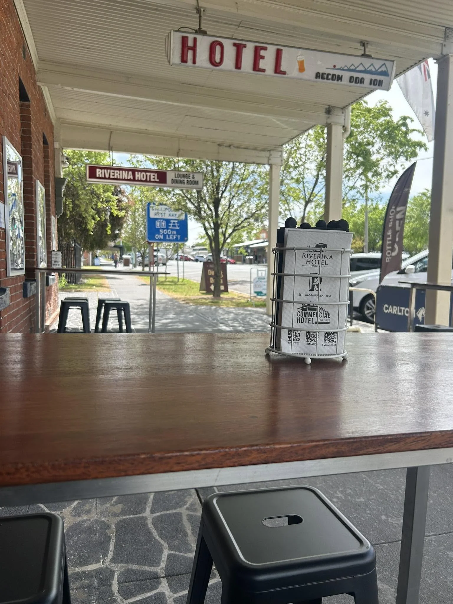 Photo taken from an outdoor sidewalk cafe or restaurant, showing black chairs and a wooden table in the foreground, with a view of a street with parked cars, trees, and business signs for a hotel, including a large sign hanging above, and a smaller s