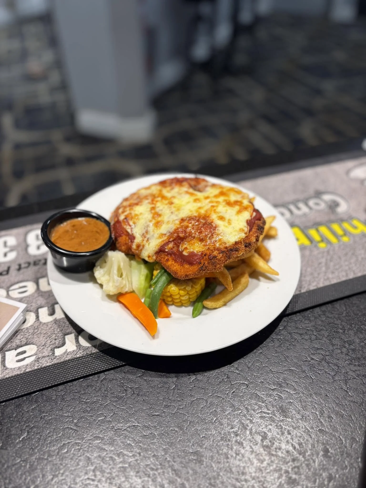 Plate of baked spaghetti with melted cheese, served with a side of vegetables including carrots, green beans, and corn, and a small cup of sauce.