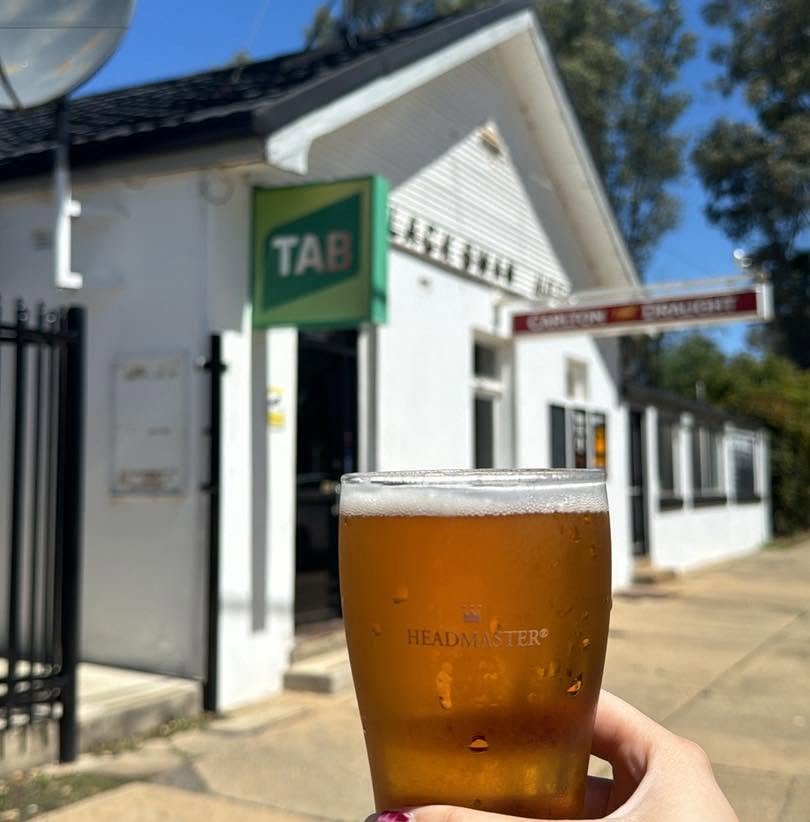 Person holding a glass of beer outside a building with a green TAB sign and a red sign, under clear blue sky.