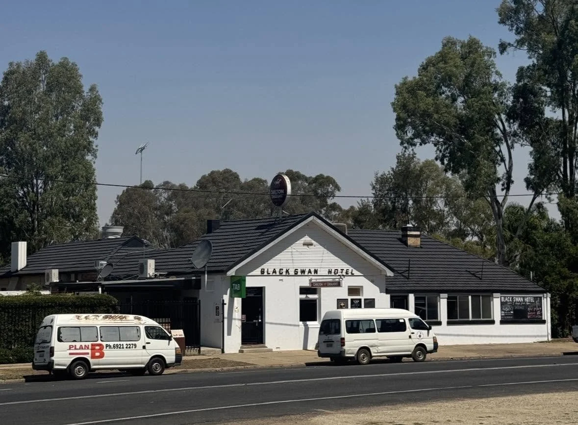 A white building with a sign that reads 'Black Swan Hotel' on the front, along a road with parked vans in front. Tall trees are in the background.