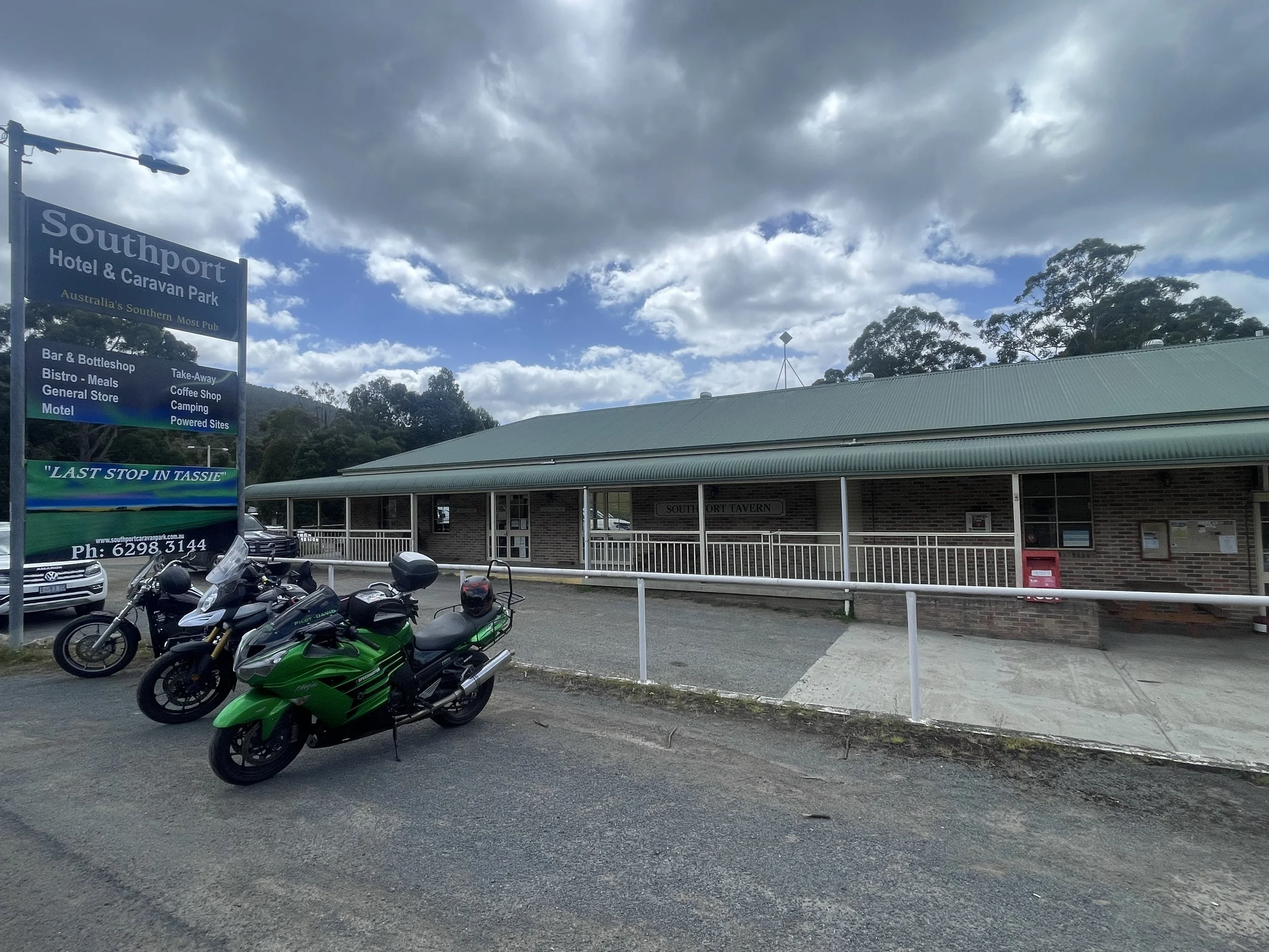 A parking lot with several motorcycles parked in the foreground, a building with a green roof, and a large sign for Southport Hotel & Caravan Park, with cloudy skies above.
