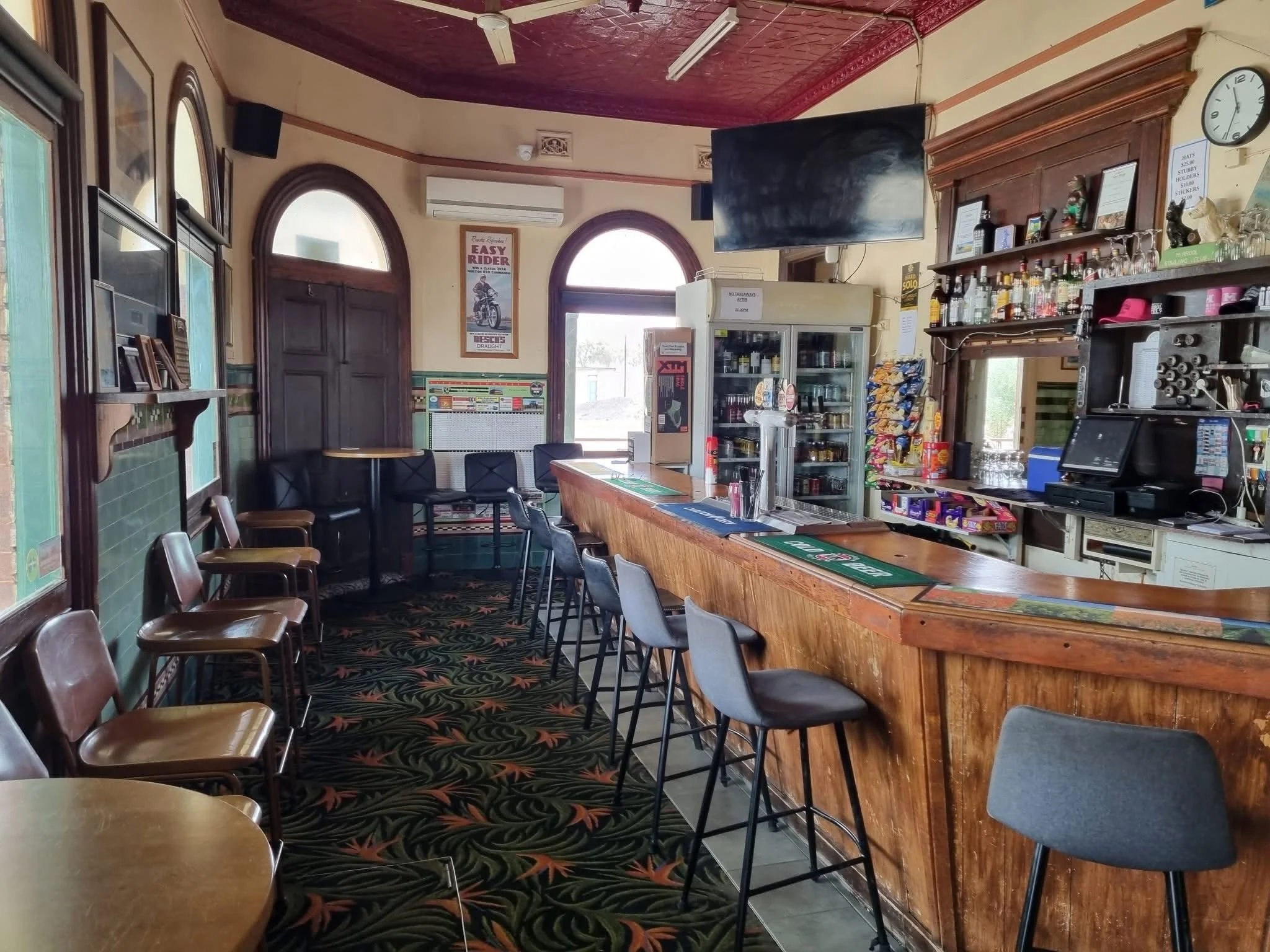 Interior of a casual bar or café with a wooden counter, bar stools, chairs, and a wall-mounted TV. Decor includes vintage posters, a clock, and a shelf with liquor bottles, snacks, and miscellaneous items. The ceiling has decorative red panels. Natur