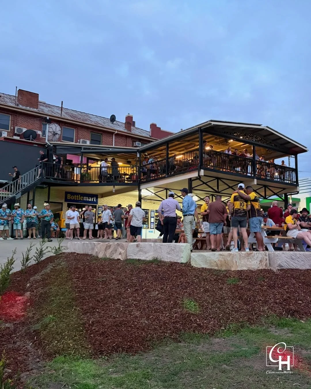 A two-story outdoor bar or restaurant with people socializing on the upper patio and in line at the lower counter. There are picnic tables on the ground level with people sitting. The building is next to an older brick structure, and the sky is cloud