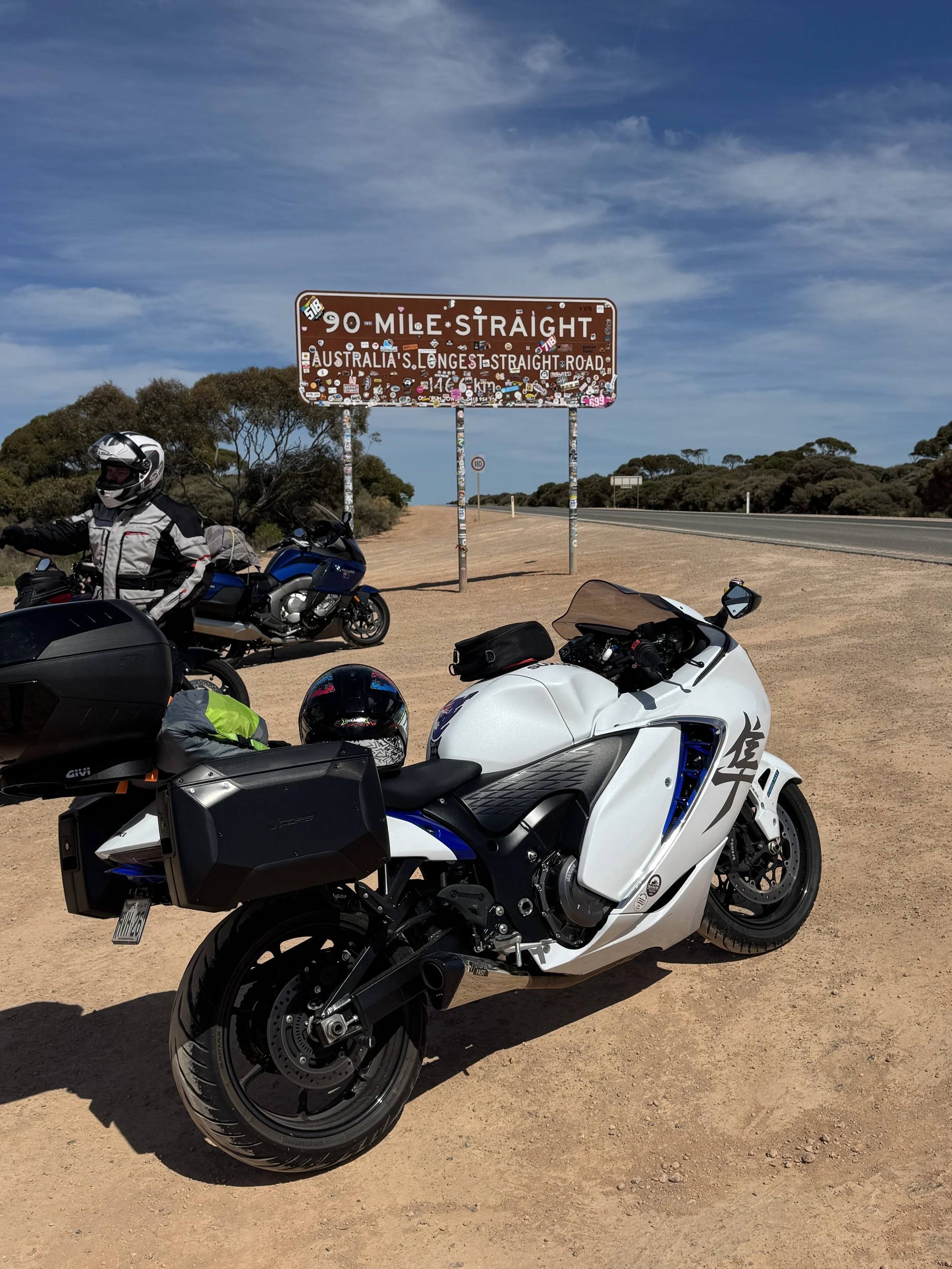A white motorcycle with black accents, parked on a dirt roadside with a helmet on the seat, and a motorcycle rider wearing a helmet and riding gear standing next to a blue motorcycle in front. There is a road sign indicating 90 miles straight, Austra