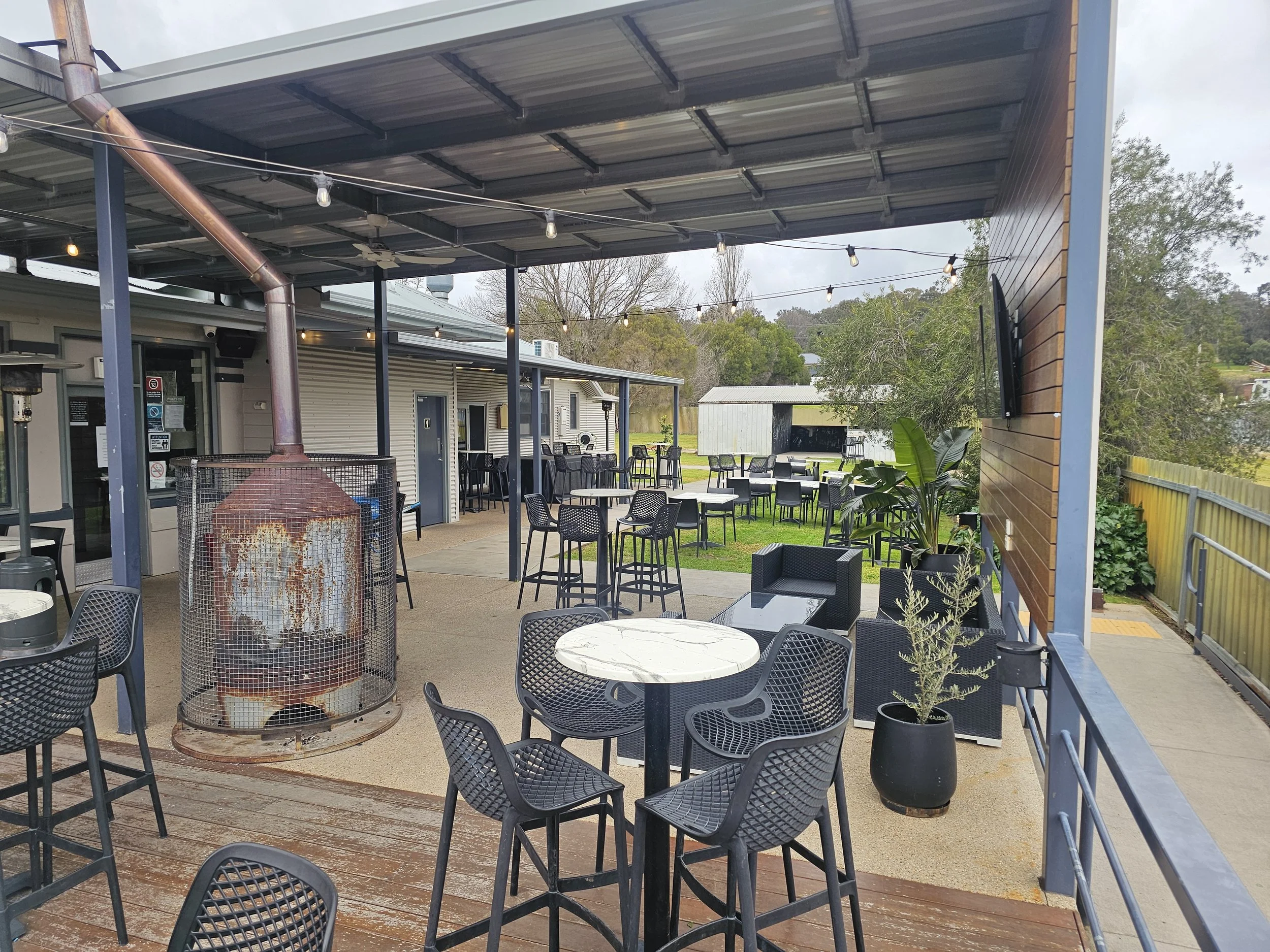 Outdoor patio area with black metal tables and chairs under a covered roof, string lights hanging, a central metal fireplace, surrounding plants, and trees in the background.
