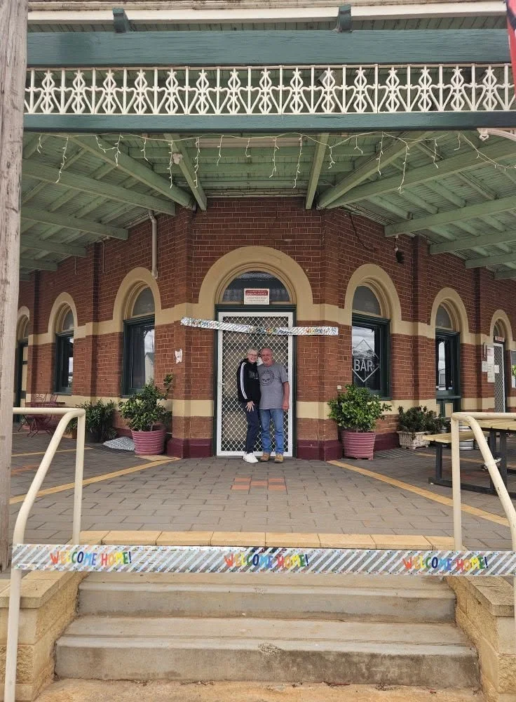 Two people standing on a small porch in front of a brick building with arched windows, decorated with Christmas lights and a "Welcome Home" banner.