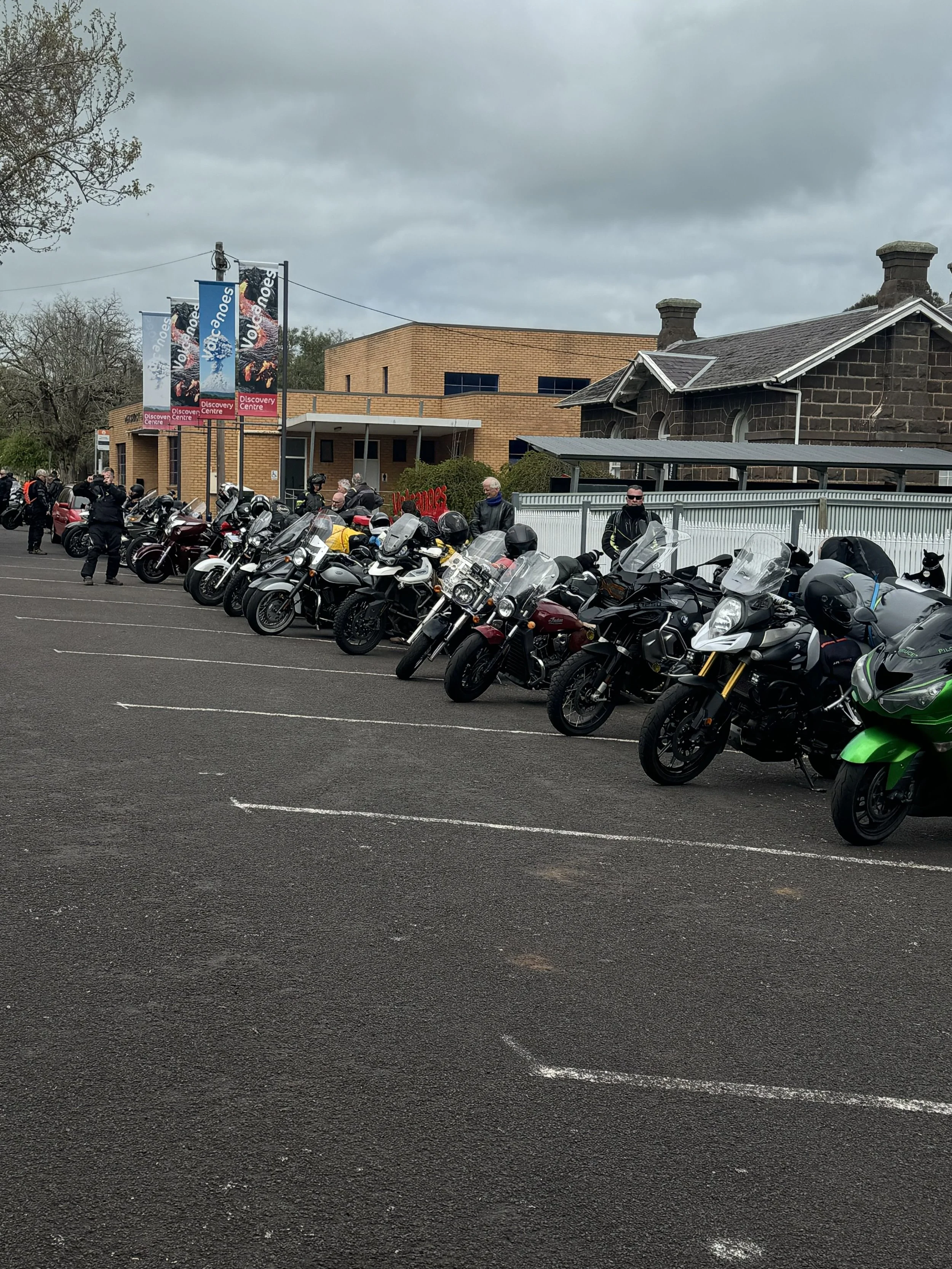 Parking lot filled with motorcycles in front of a commercial building with banners that read 'Voyages' and 'Discover Centre'. Several people are gathered around the bikes, and the background shows a cloudy sky.