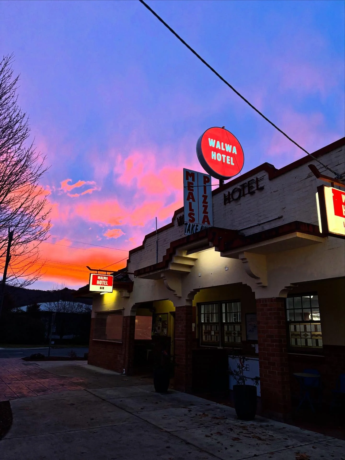 Sunset sky with pink, purple, and orange clouds behind a building with illuminated signs for Walwa Hotel, bar, pizza, and meals. The building has brick and white exterior walls with windows and decorative arches.