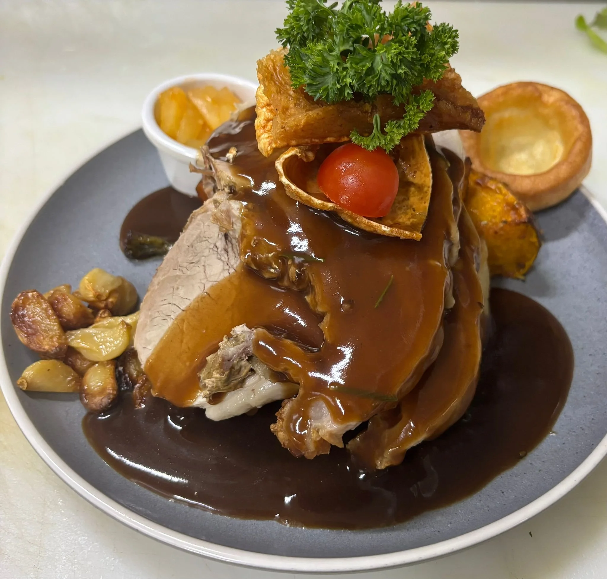 A plate with sliced meat topped with brown gravy, garnished with parsley and cherry tomato, with side dishes of fried potato wedges and baked potato halves in a small bowl.