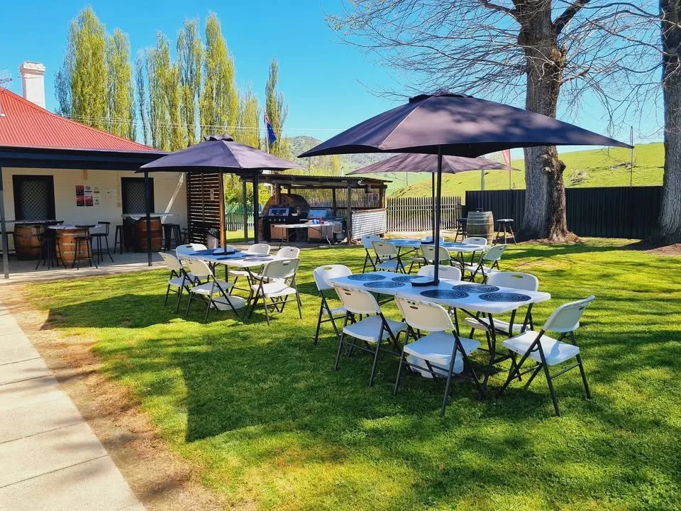 Outdoor dining area with tables, chairs, and large umbrellas on a grassy yard, with a building and trees in the background.
