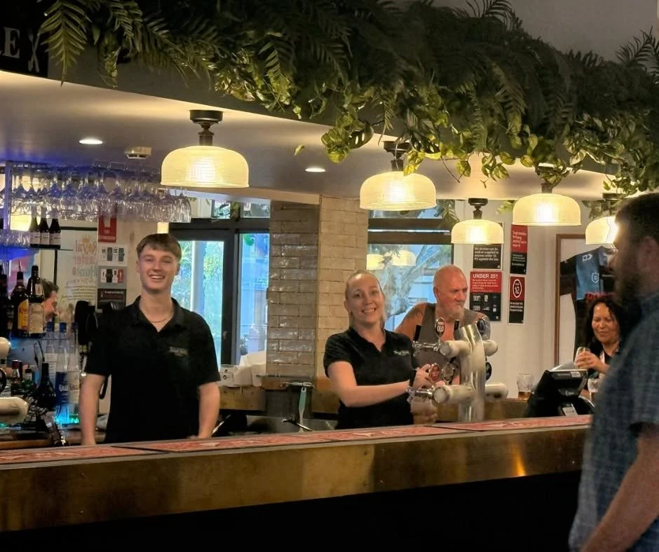 Bar with two smiling bartenders serving drinks, and a customer at the counter, decorated with hanging lights and greenery.