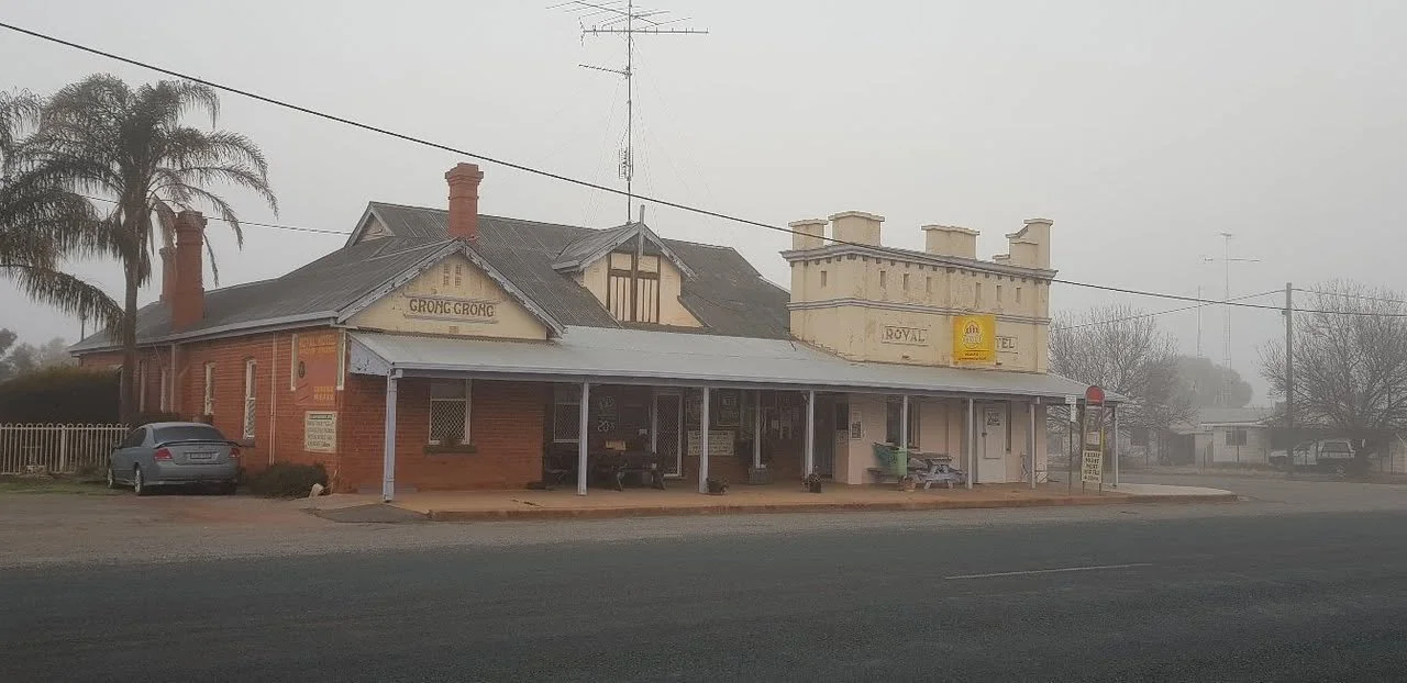 An old-style building with a brick exterior, a covered porch, and signage indicating a hotel called "Royal Hotel." There are a few trees and a parked car in front of the building, and the scene appears foggy or misty.
