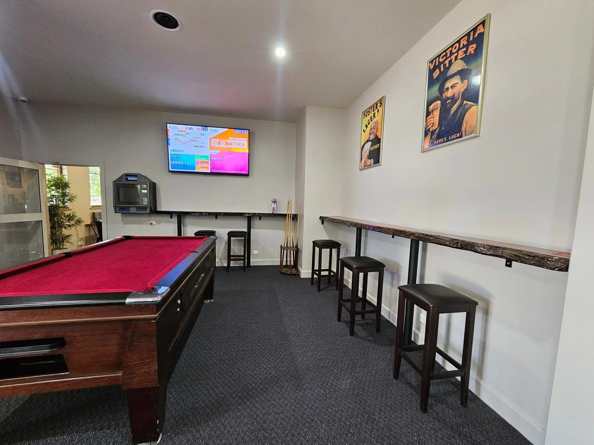 An interior room with a pool table, high chairs along the wall, a wall-mounted TV displaying lottery numbers, and posters of Victor Bitet and Foster's Lager on the wall.