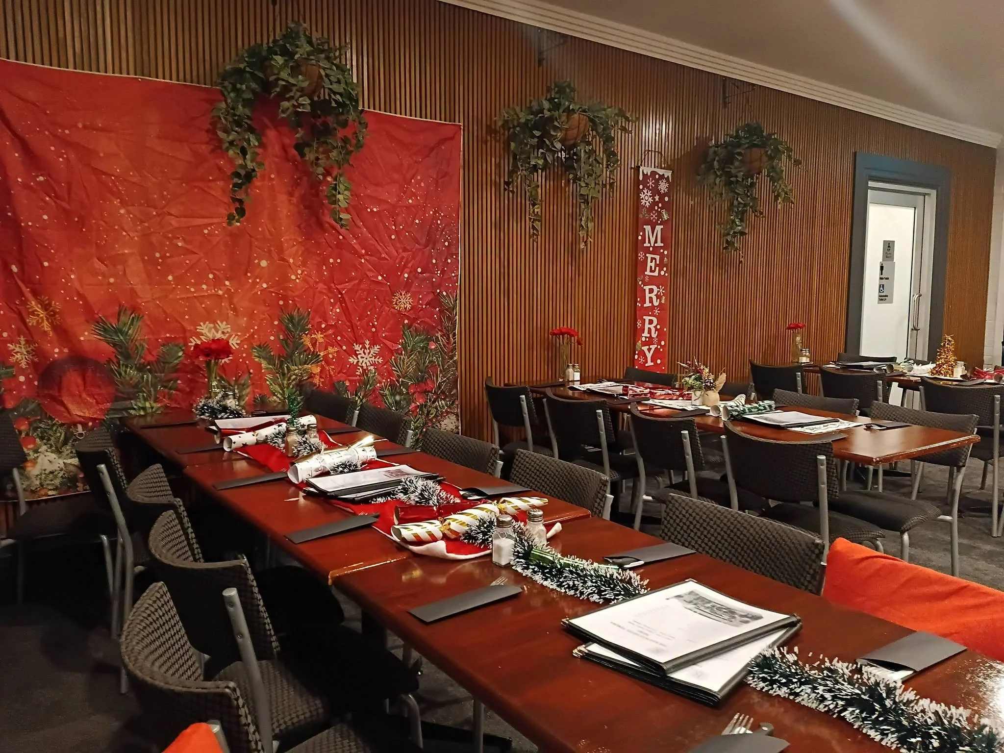 Restaurant or event hall decorated for Christmas with a red backdrop, green garlands, and holiday-themed table settings.