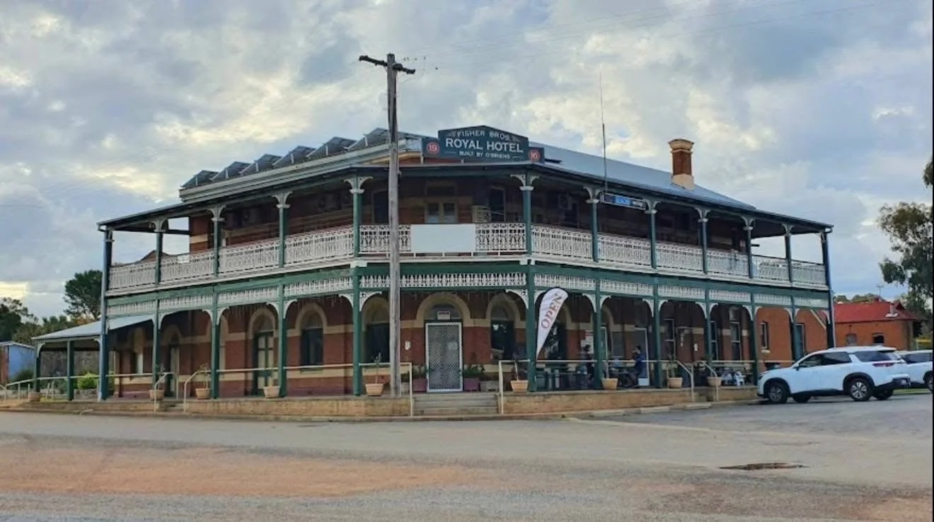 Historic two-story brick hotel with a large upper balcony and decorative columns, labeled 'Royal Hotel' with a sign for reservations, parked cars in front, and a cloudy sky.