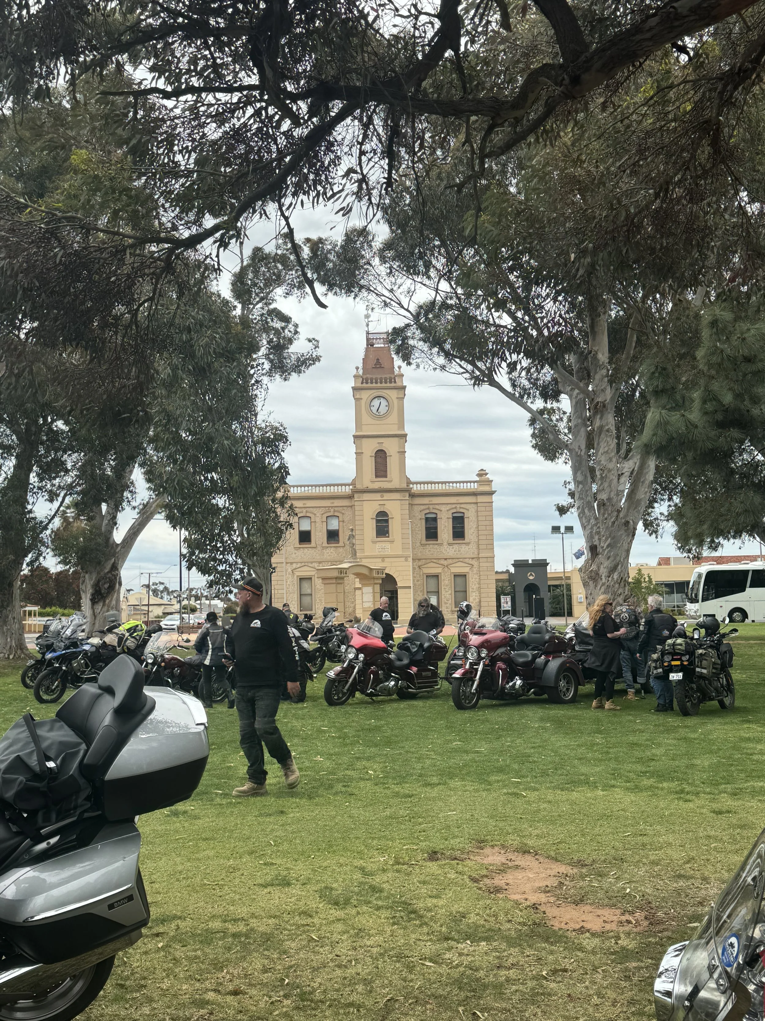 A historic yellow building with a clock tower, surrounded by trees and people with motorcycles in a park.