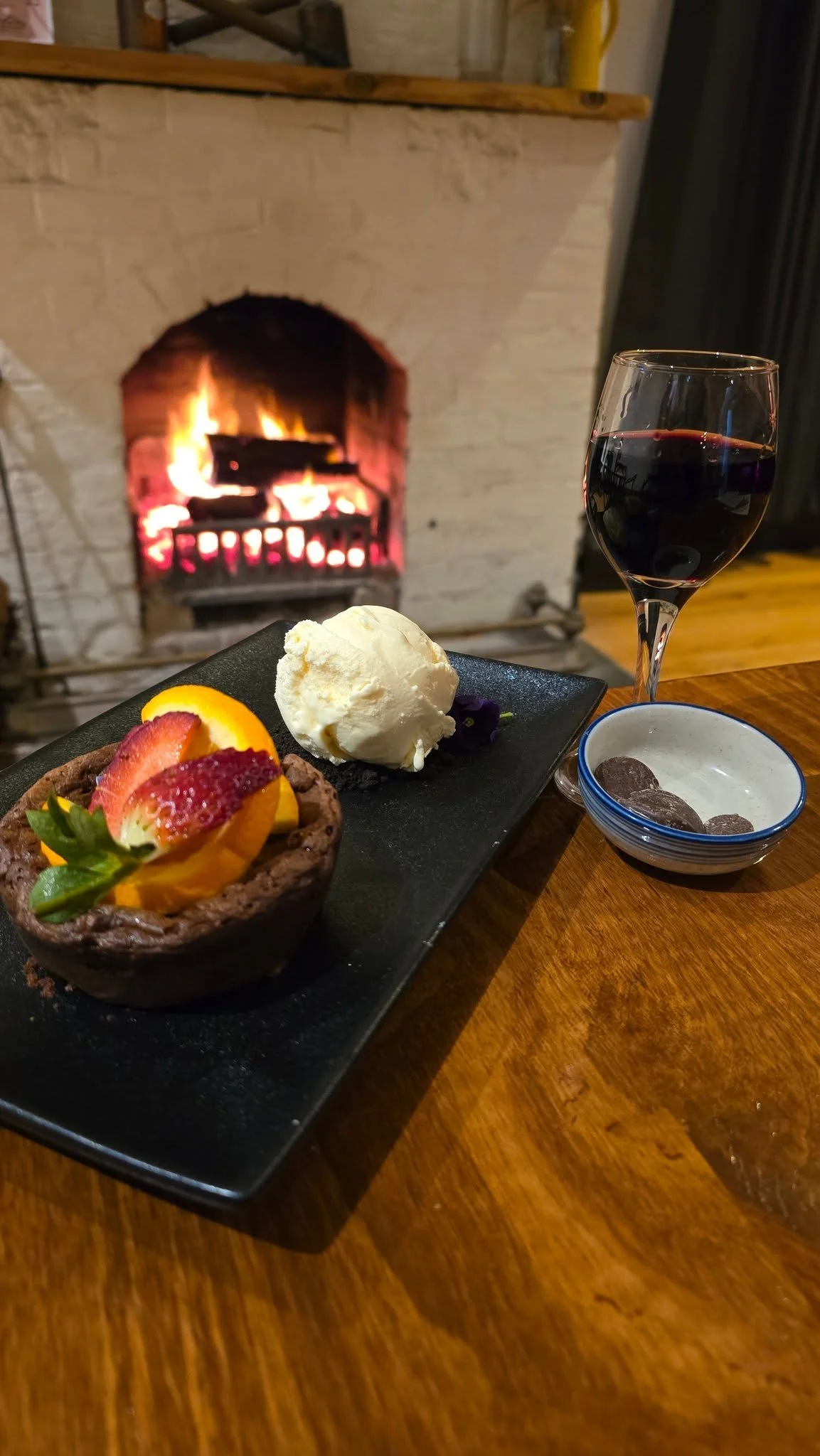 A dessert plate with a chocolate tart topped with sliced fruits, a scoop of vanilla ice cream, a glass of red wine, and a small bowl of chocolate candies in front of a fireplace.