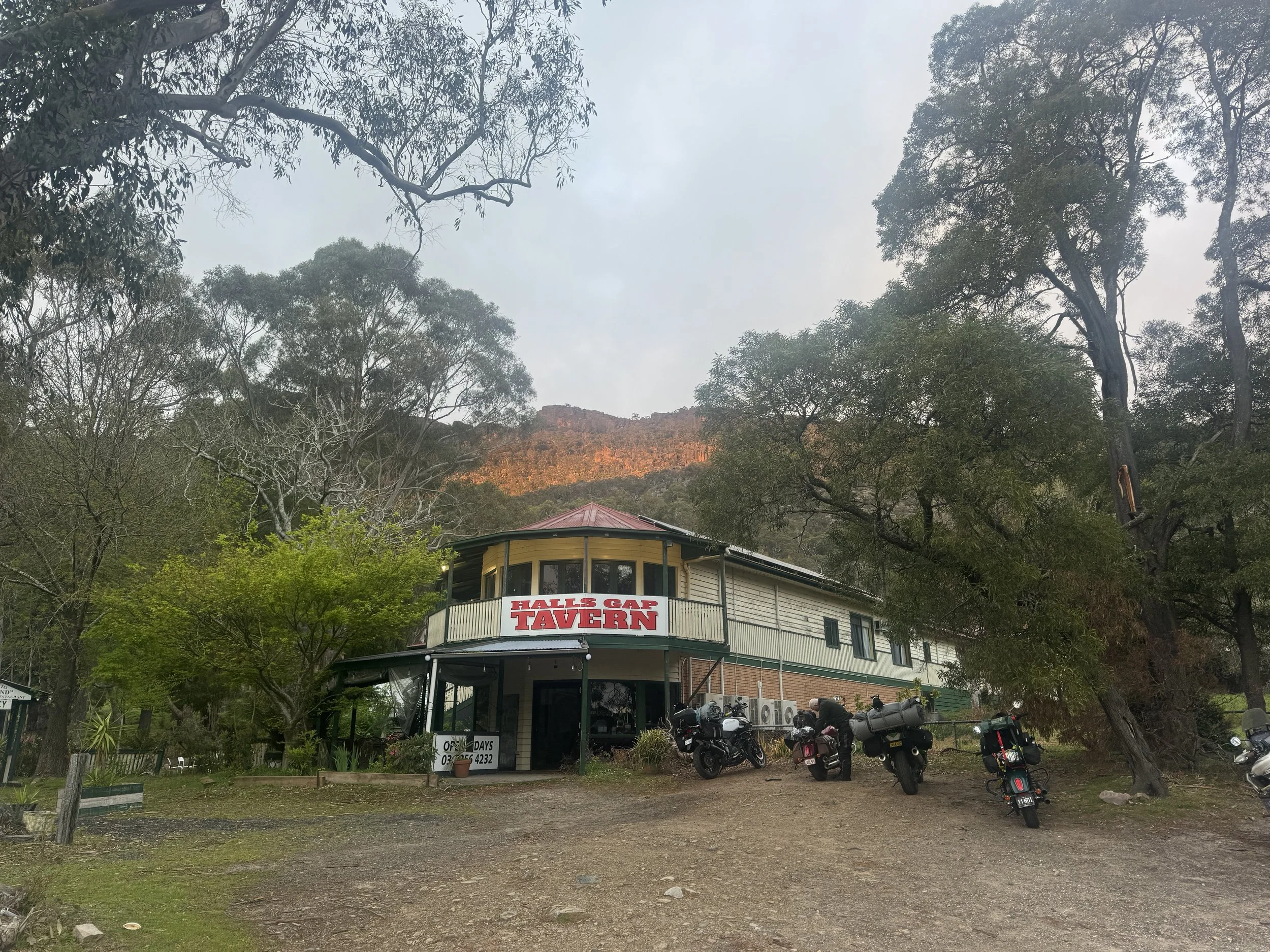 A building with a sign that reads "Halls Gap Tavern" located among trees with rugged hills in the background, several motorcycles parked in front and a dirt ground.
