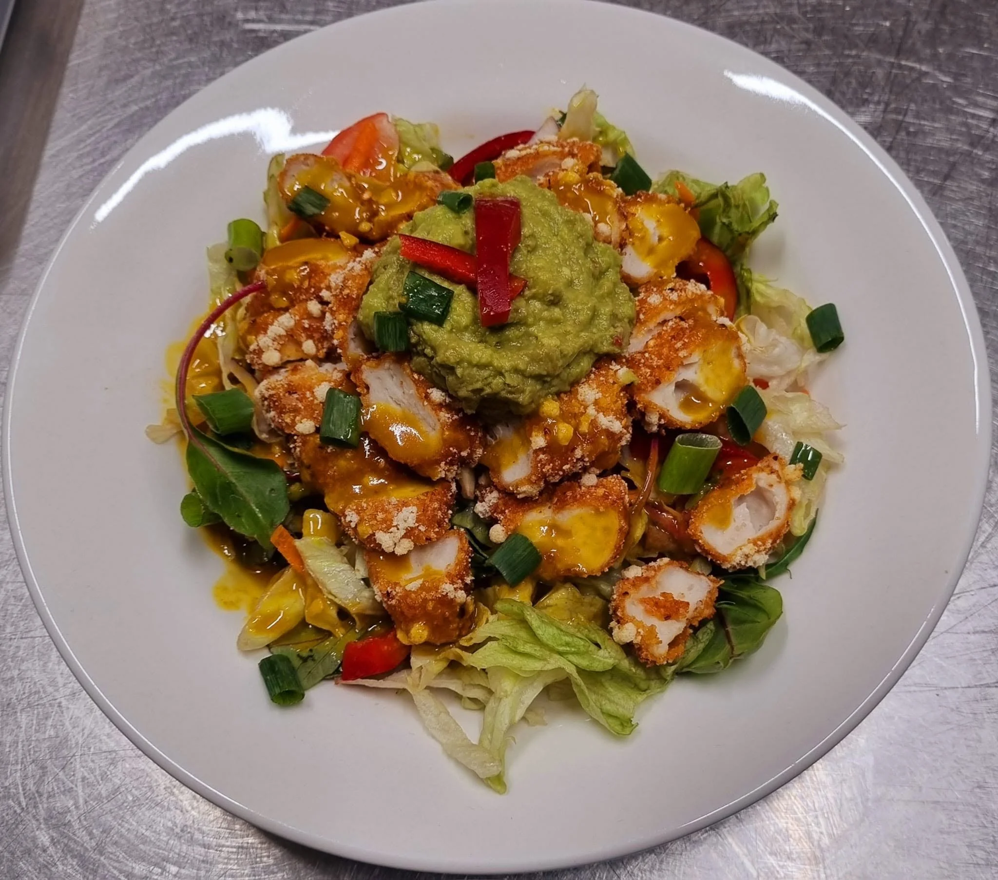 A white plate with breaded fish fillets topped with guacamole, chopped green onions, and a yellow sauce, served over chopped lettuce with sliced tomatoes and carrots.