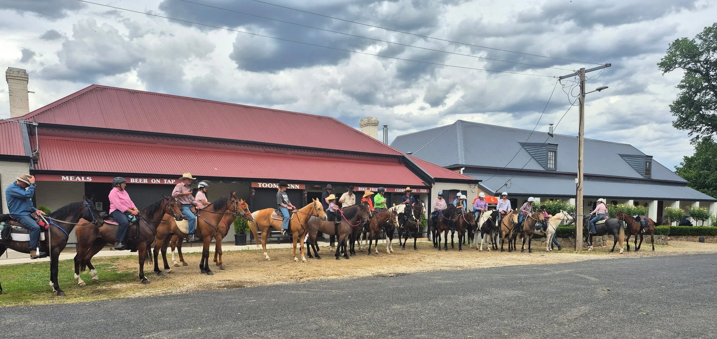 Group of people riding horses in front of a building with a red roof and a sign that says 'Meals, Beer on Tap, Toom Inn, Bike Friendly, Closed on Tue'.