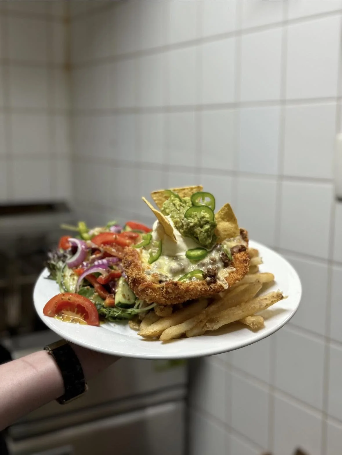 Plate of food with fried chicken topped with jalapeños, sour cream, and chips, served with French fries and a side salad with tomatoes, cucumbers, and red onions.