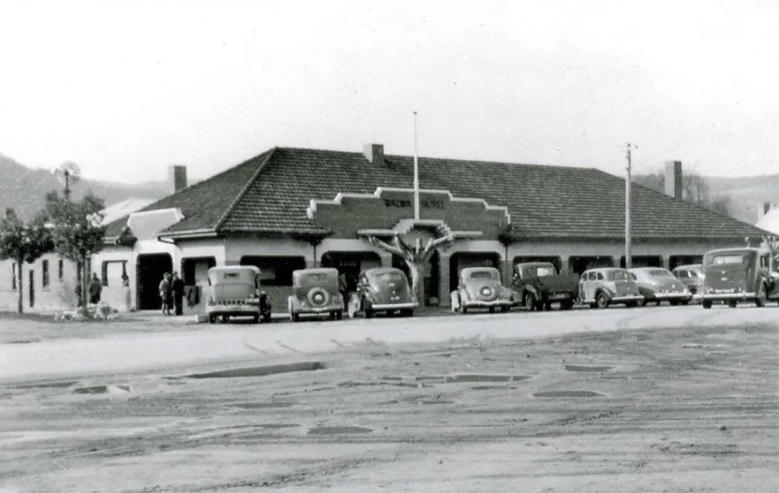 A black and white photo of a small building labeled "ELBURN HOTEL" with vintage cars parked in front of it, and a few people standing near the entrance.