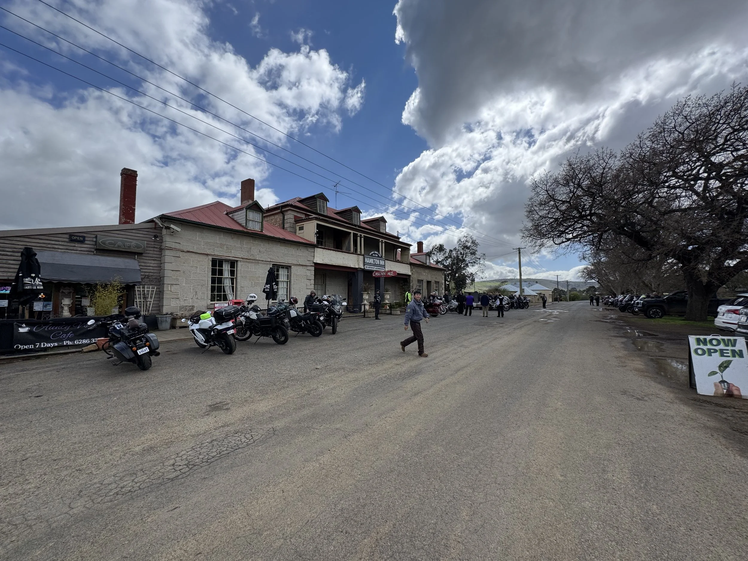 A street scene featuring a historic building with a red roof and multiple chimneys, students and motorcyclists gathered outside, trees with no leaves, parked cars, and a partly cloudy sky.